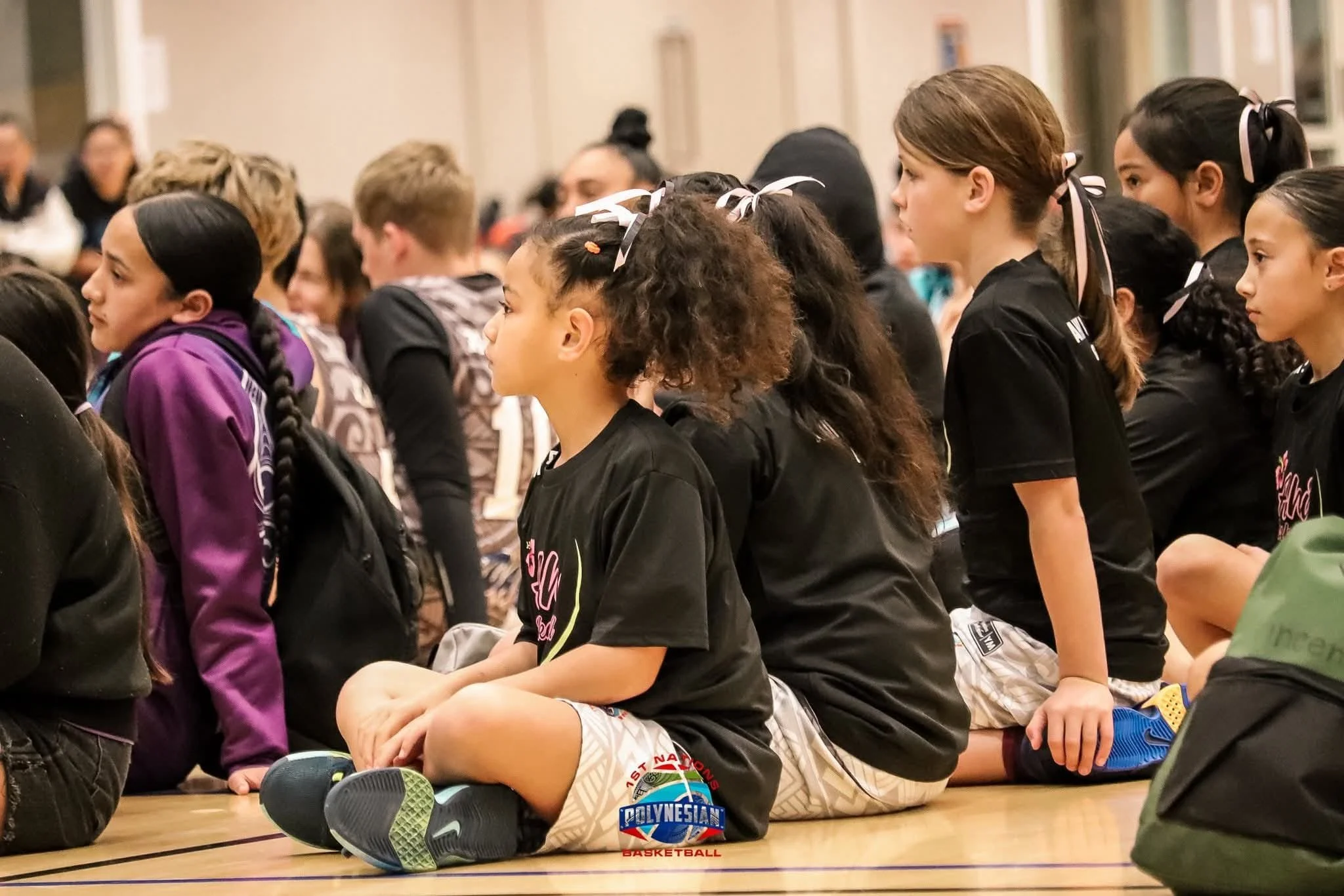 A group of children sitting cross-legged on the floor, paying attention at an indoor event or assembly. Many children have ribbons in their hair and are wearing black T-shirts and athletic shorts.