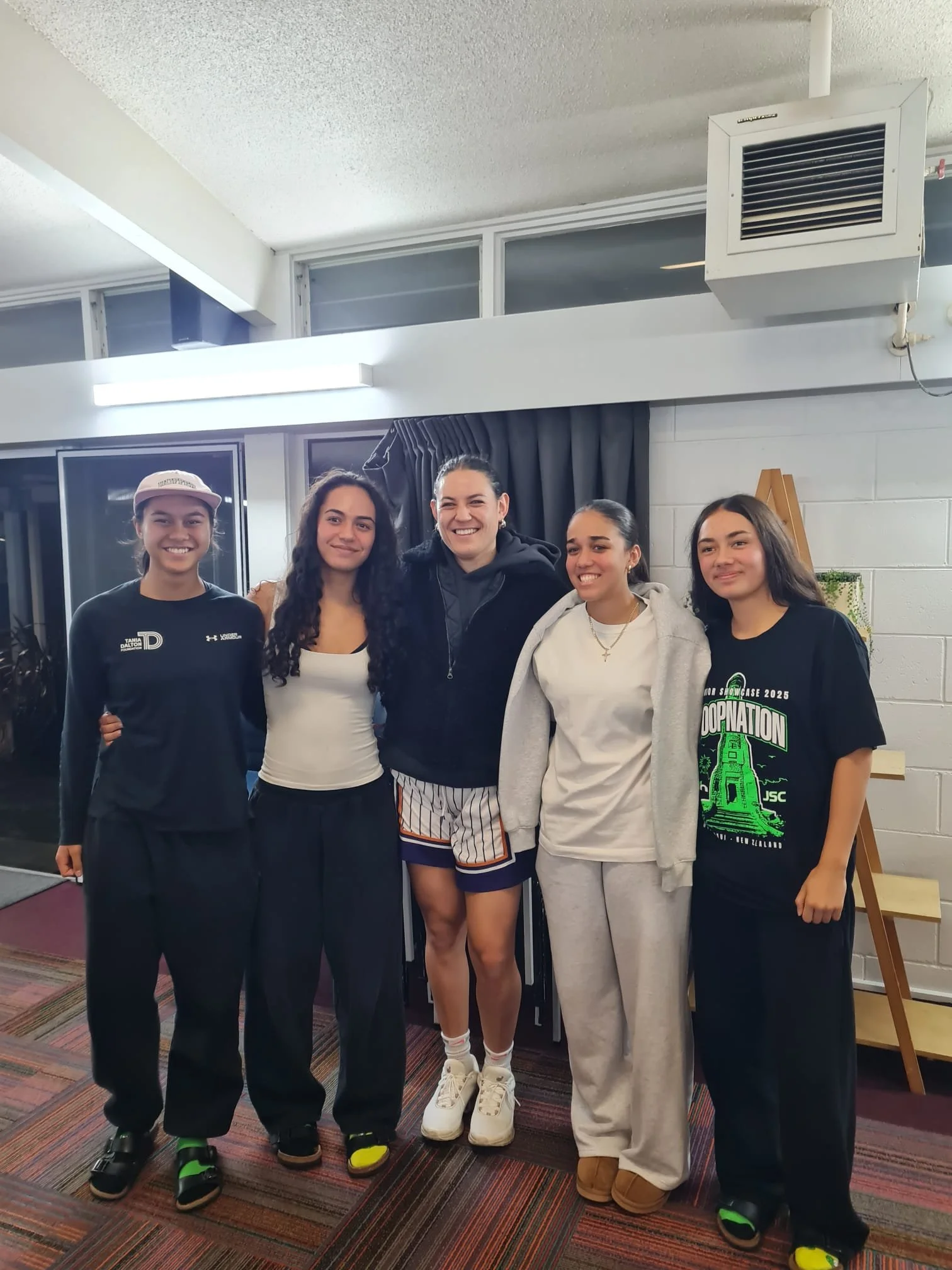 Five women standing together indoors, smiling for a photo. They are dressed casually, with some wearing sports or comfortable clothes, and sneakers. The background includes large windows, gray curtains, and a white brick wall.