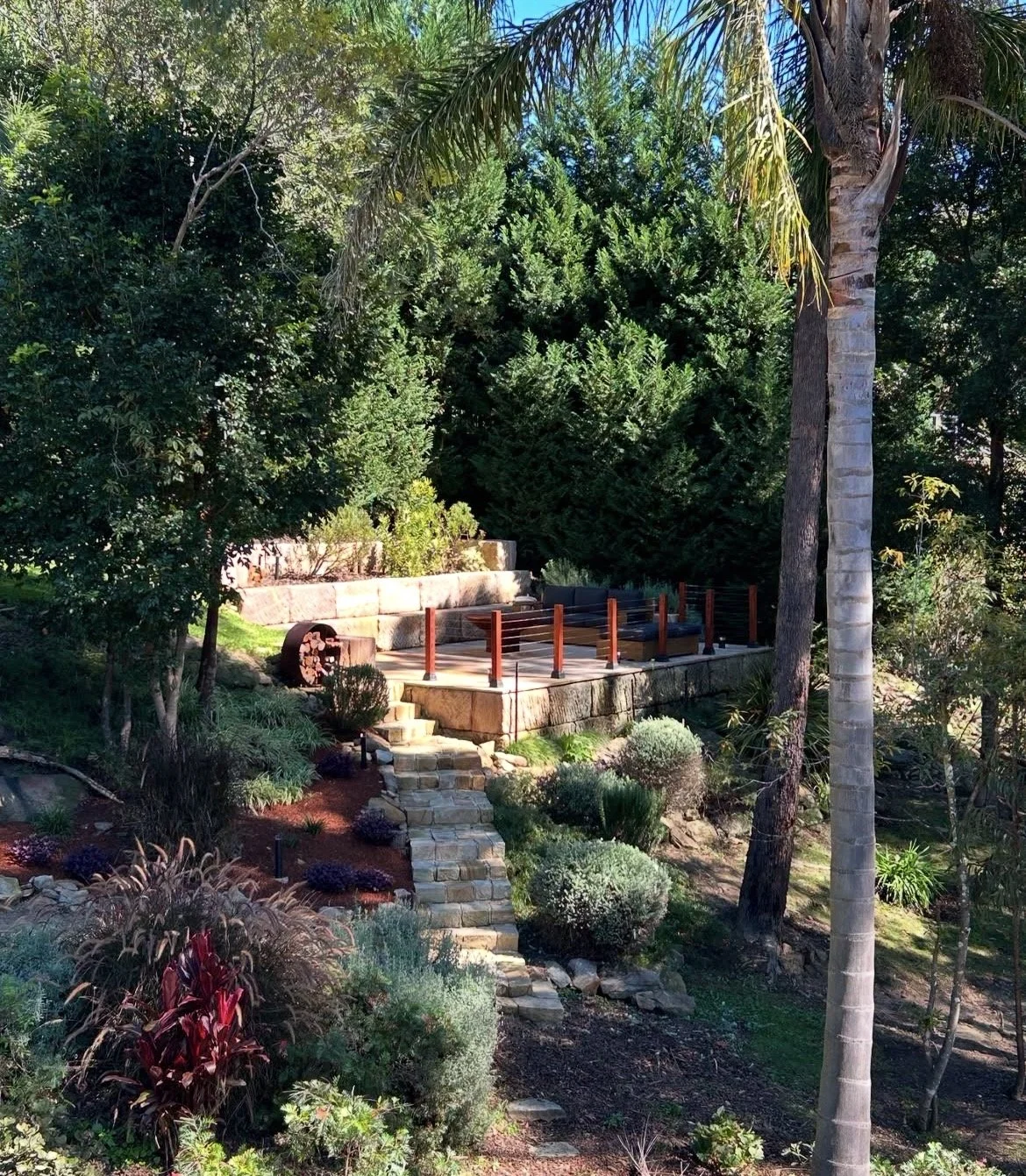 A landscaped backyard with stone steps leading up to a raised patio area with outdoor furniture, surrounded by lush green trees, bushes, and tropical plants, under a bright blue sky.