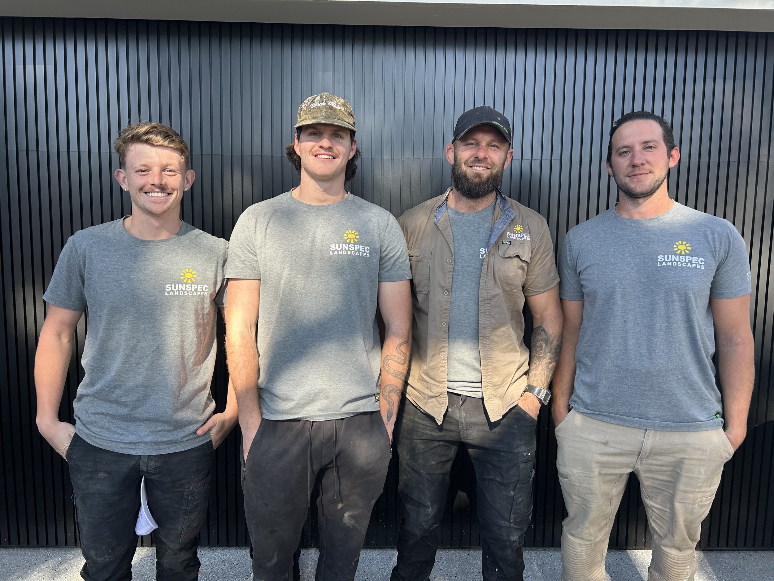 Four men standing in front of a black corrugated metal wall, wearing gray T-shirts that read 'SUNSPEC LANDSCAPES' with a yellow sun logo, smiling at the camera.