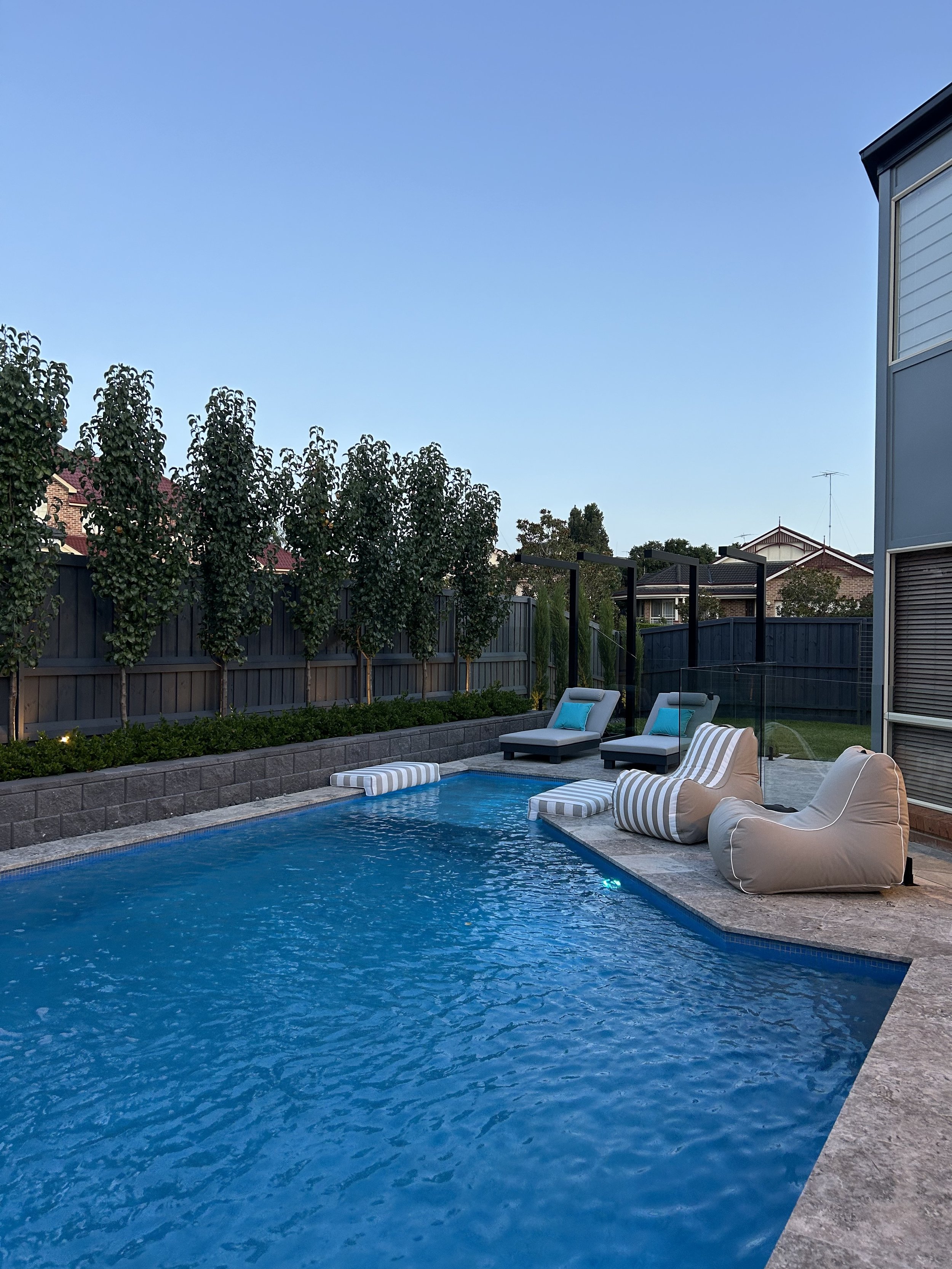 Backyard swimming pool with poolside lounge chairs, striped cushions, and bean bags, surrounded by a fence and trees, with neighboring houses and a clear sky in the background.