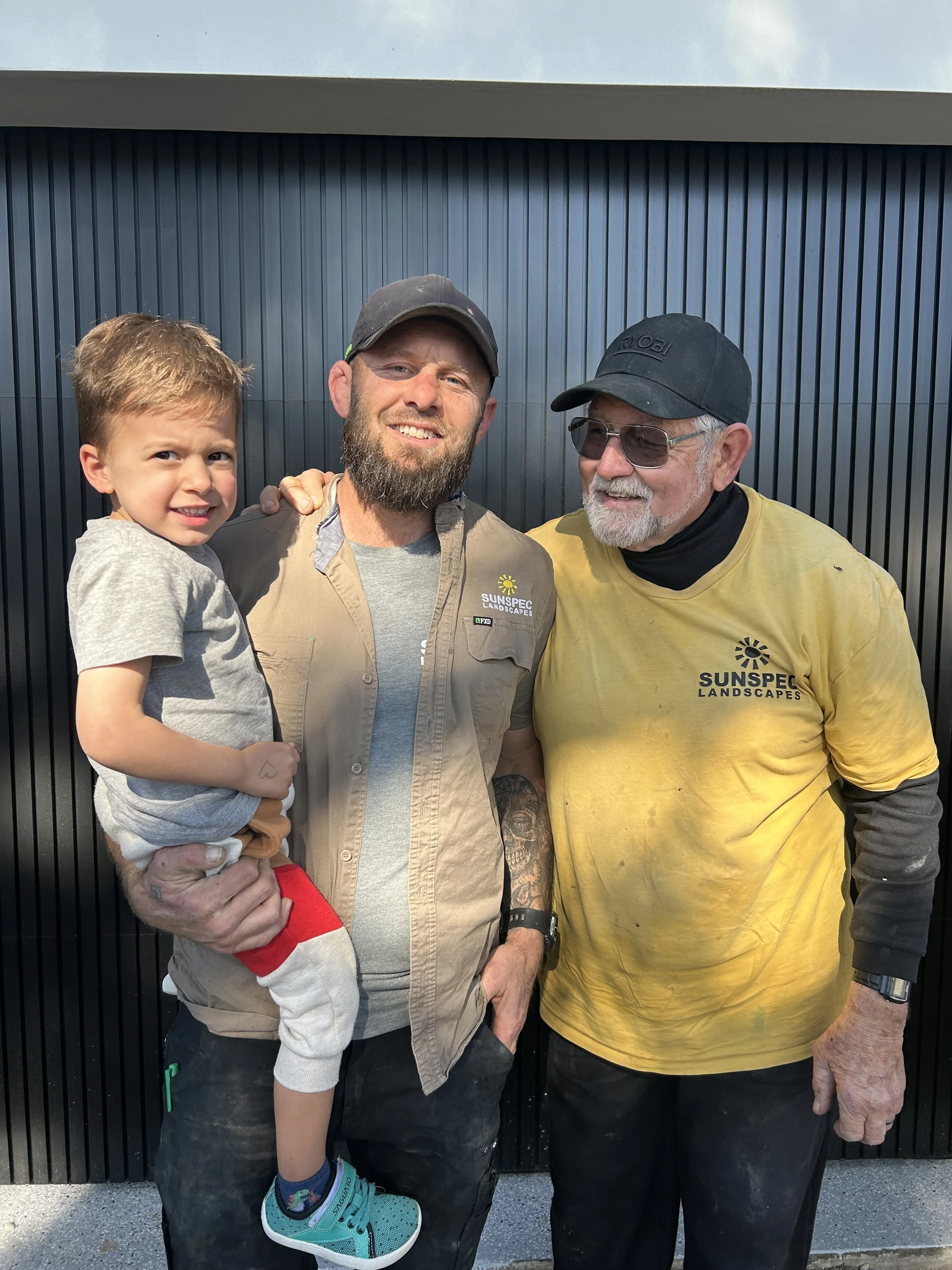 Three people standing in front of a black wall, smiling for the camera. A young boy is being held by a man with a beard and a cap, and another man in a yellow shirt with glasses is standing beside them.