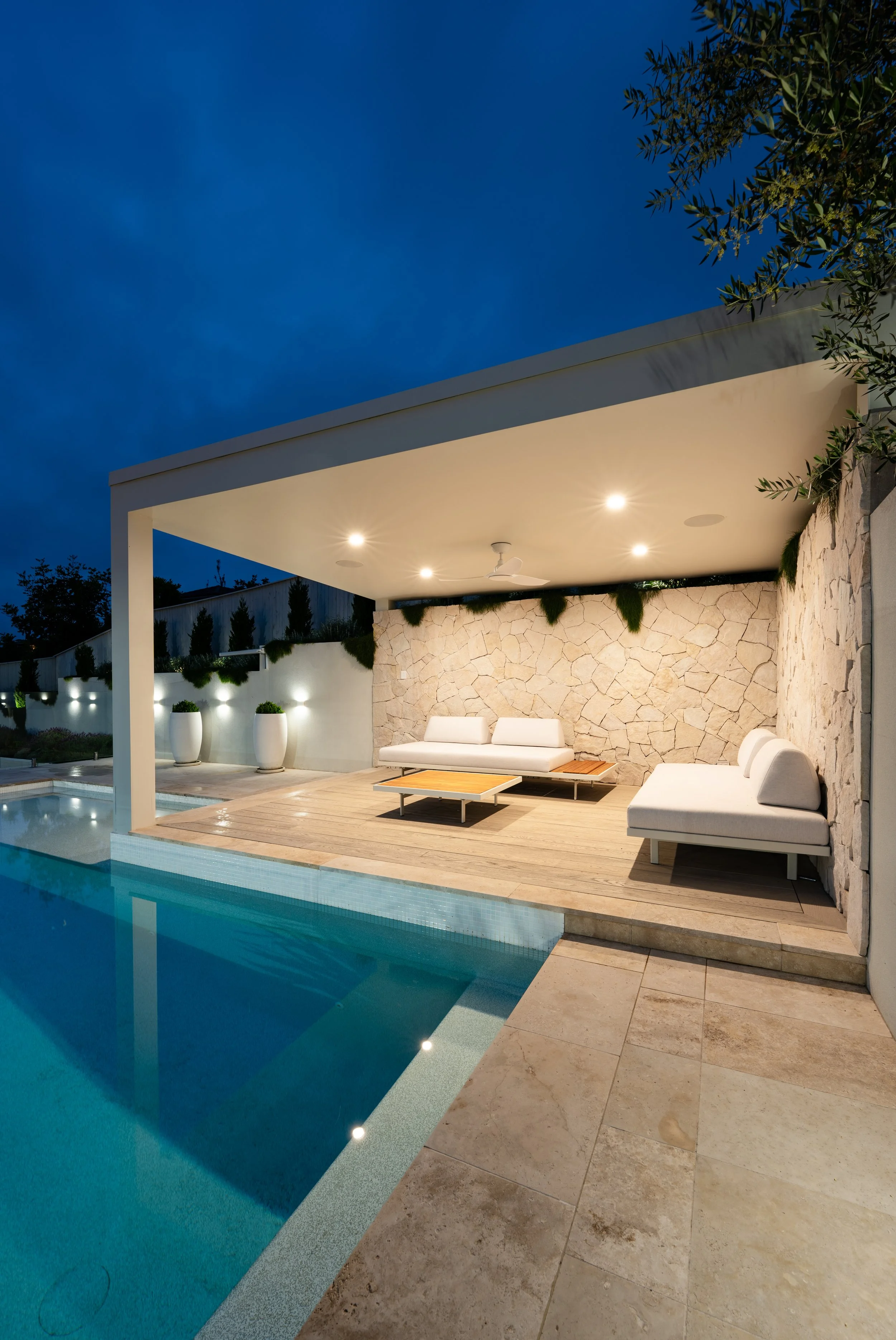 Night view of a modern outdoor patio with a swimming pool, white outdoor furniture, stone wall, and warm ceiling lights under a covered area.