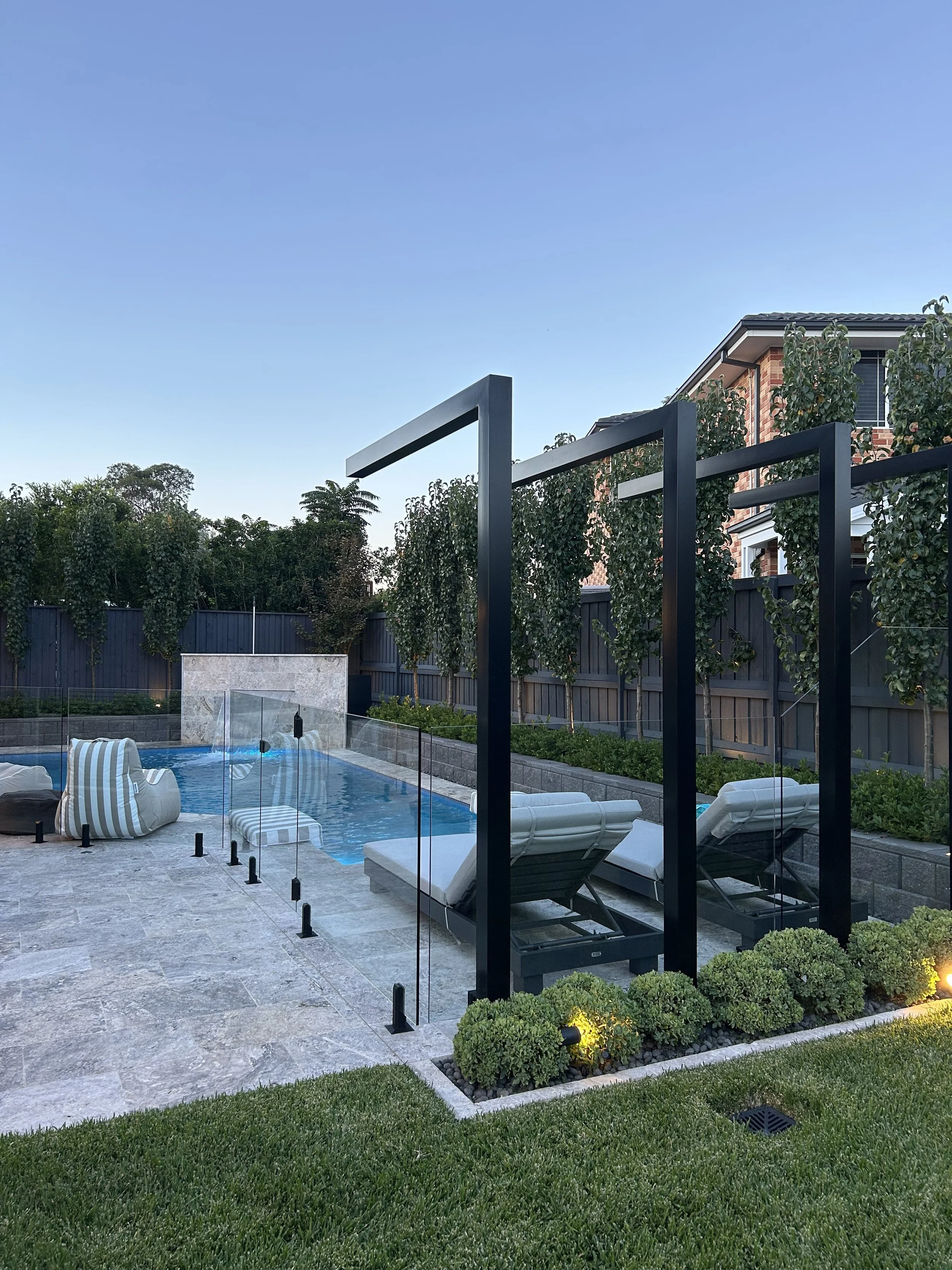 A backyard with a swimming pool, lounge chairs, a modern glass fence, green bushes, and trees, with a house in the background under a clear blue sky.
