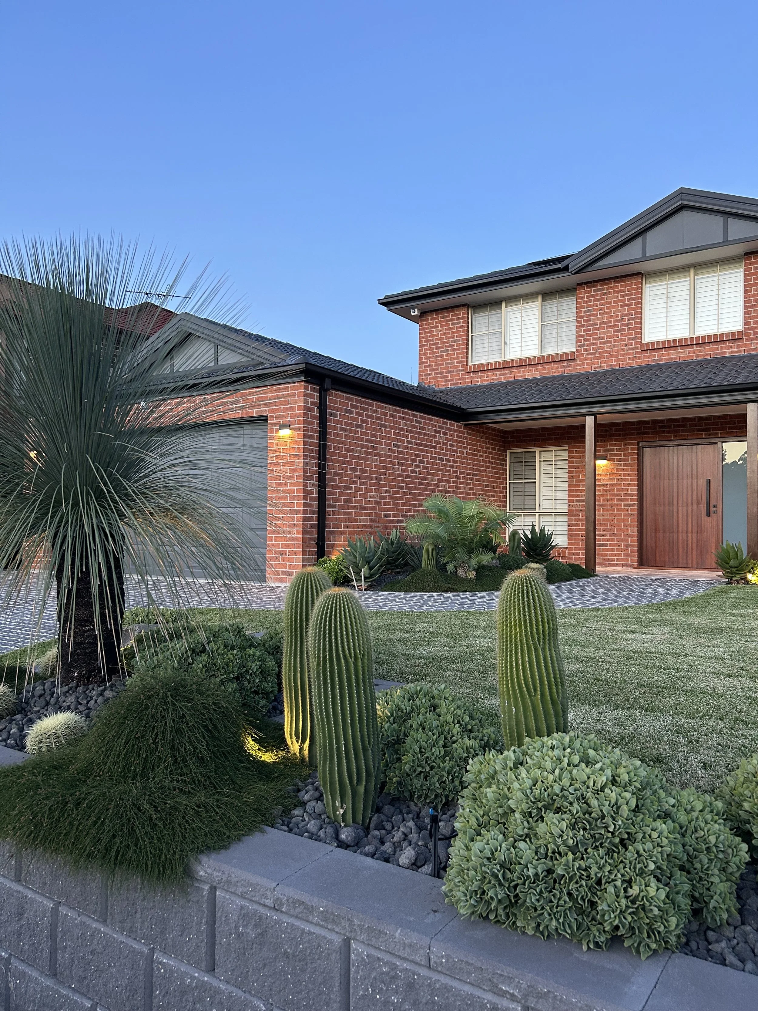 Front yard of a brick house with desert landscaping, including tall cacti, small palms, and bushes, with a paved walkway and gray brick border.