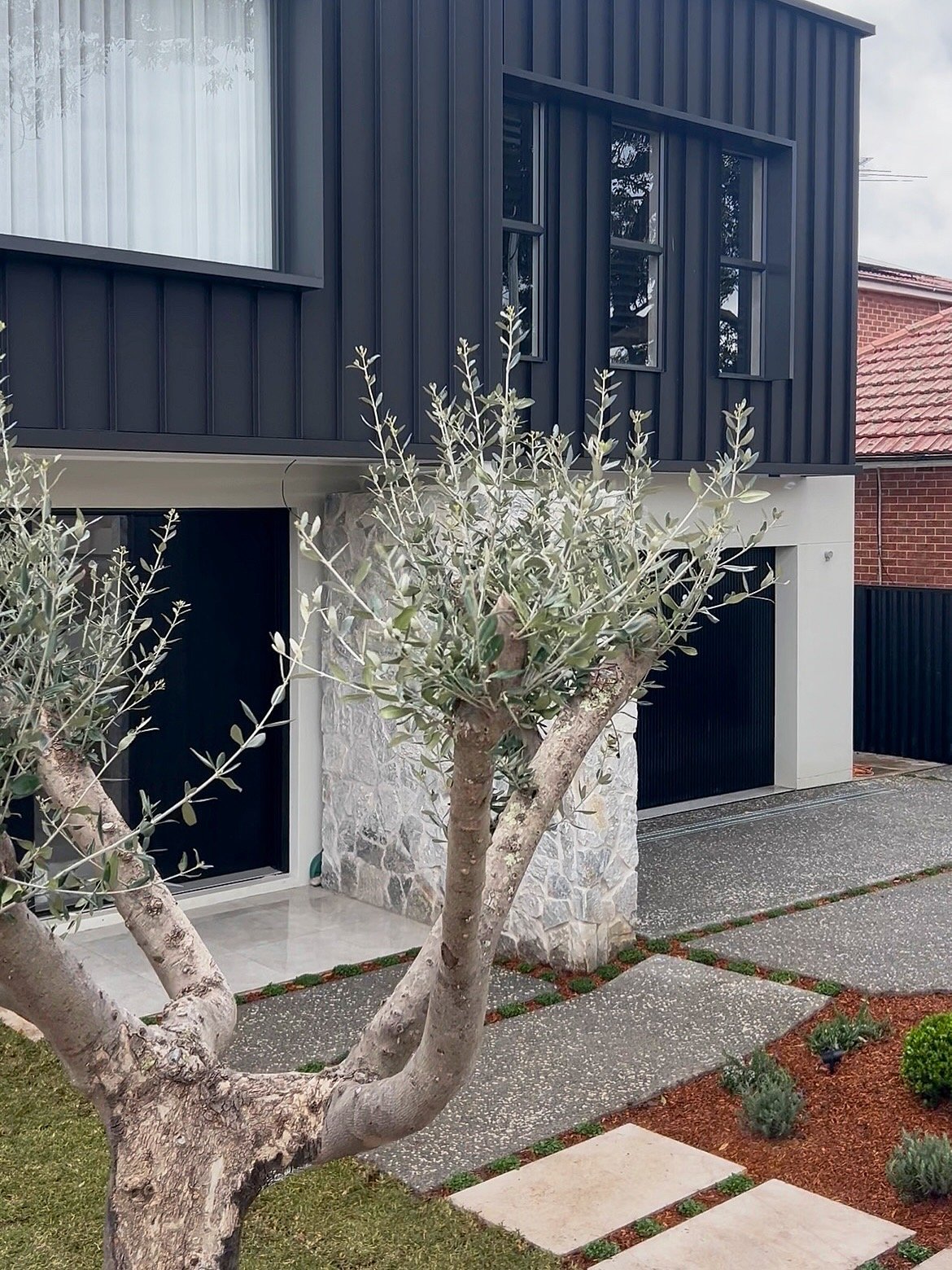 Modern two-story house with dark metal siding, stone accents, and large windows, surrounded by a landscaped yard with a tree, plants, and a pathway.