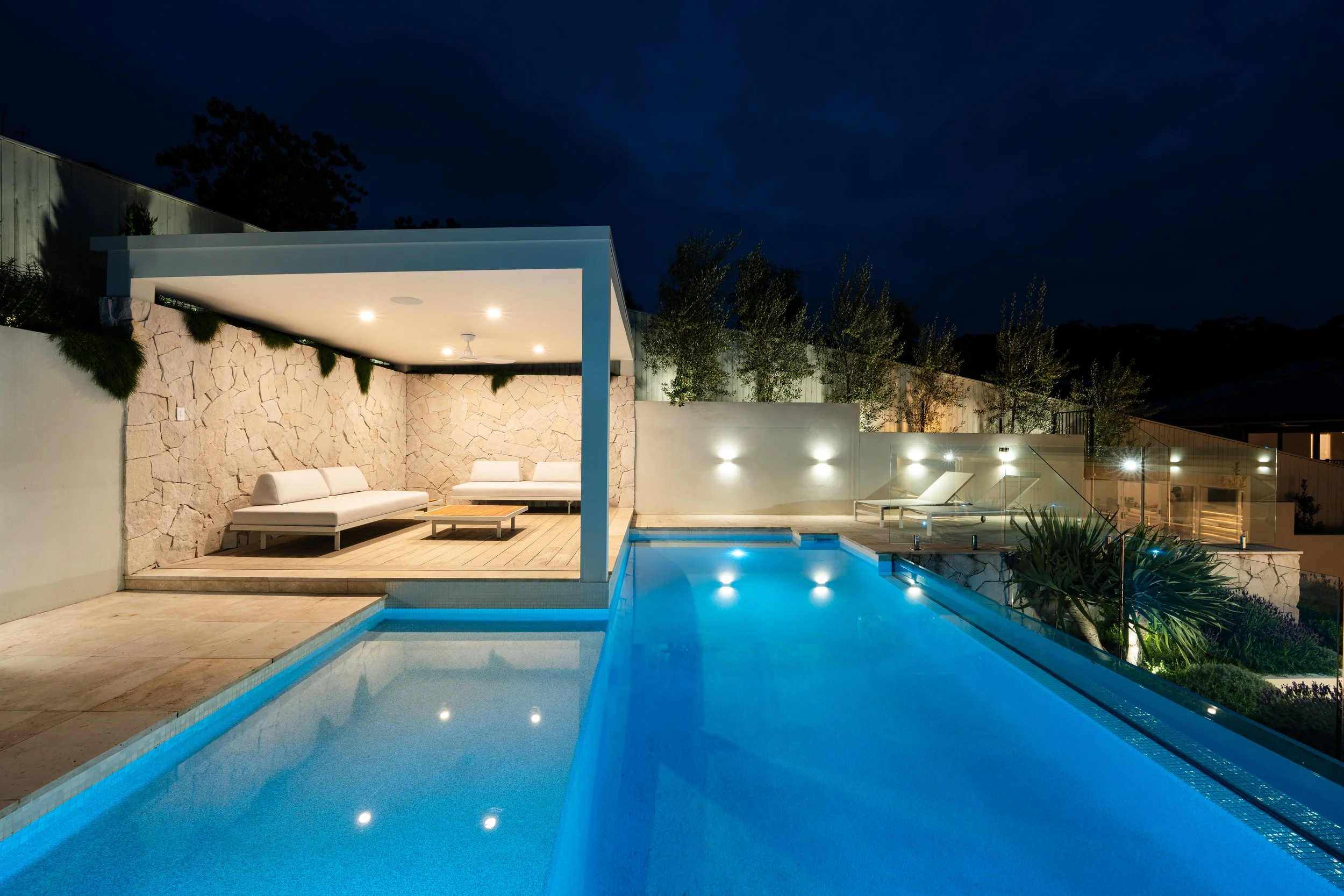 Night view of a modern backyard with a swimming pool, outdoor seating area with white couches, stone wall, and landscaped trees, illuminated by outdoor lights.