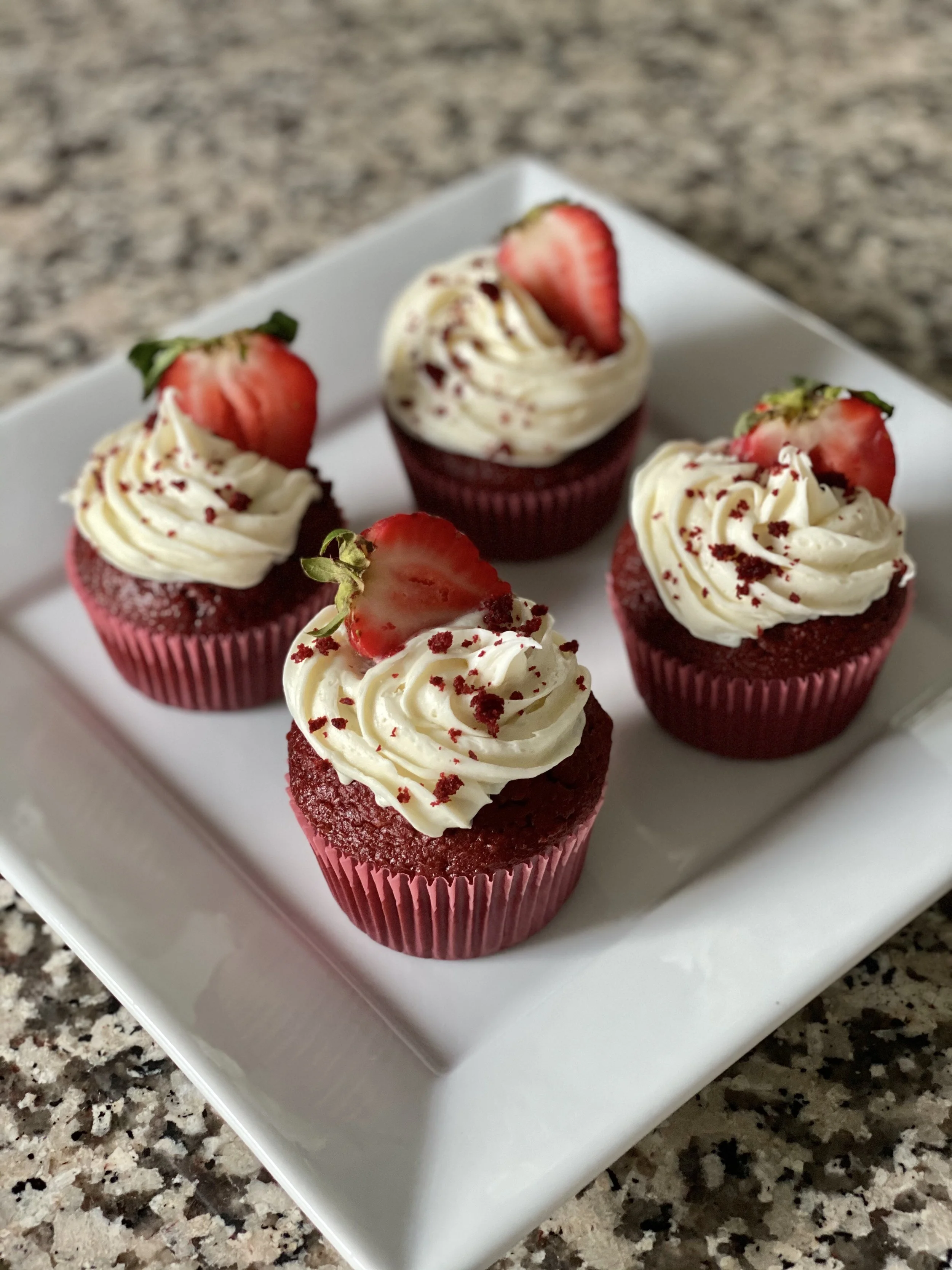 Four red velvet cupcakes with white frosting, topped with sliced strawberries and red sprinkles, arranged on a white square plate on a speckled countertop.