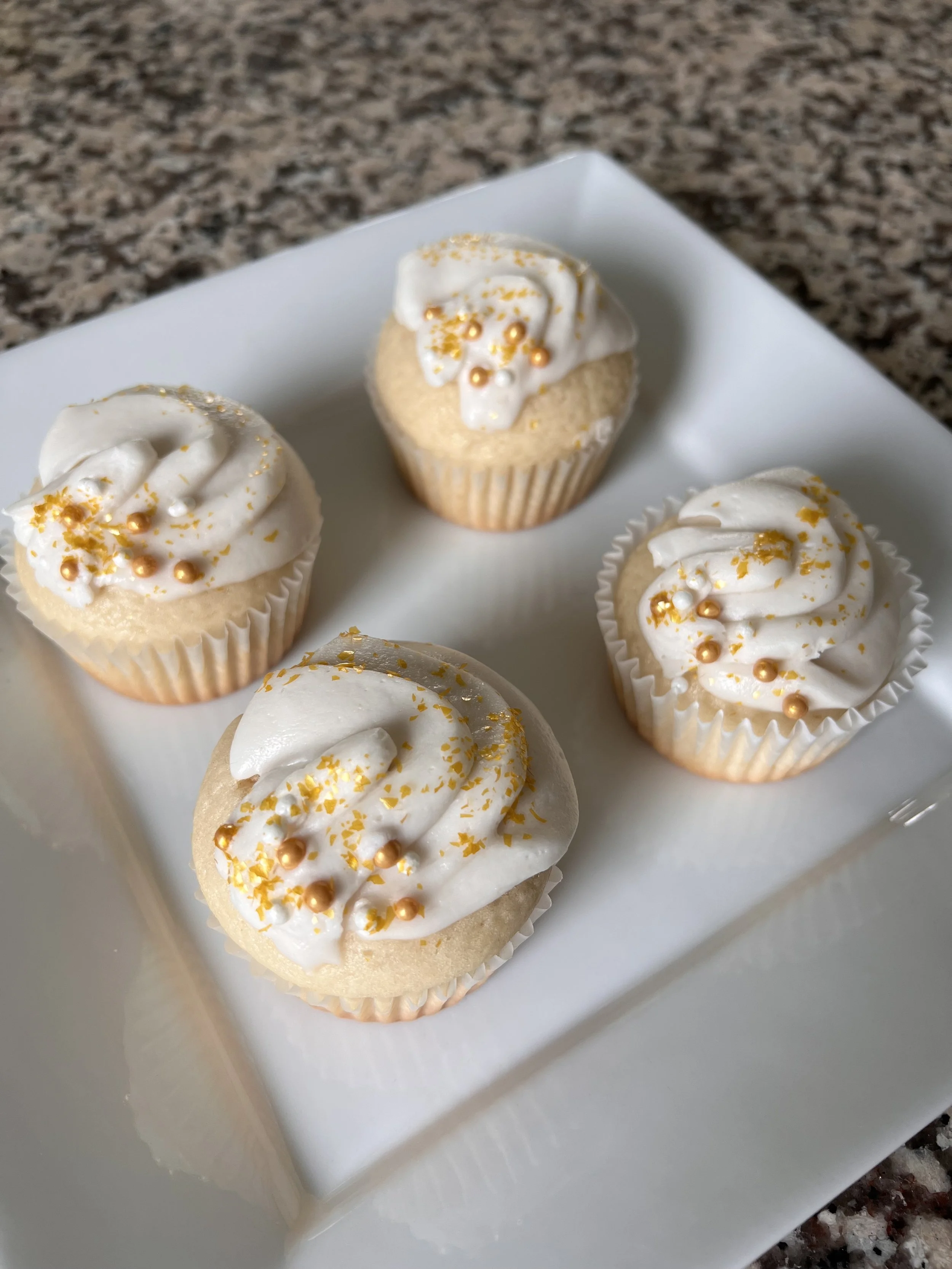 Four vegan small vanilla cupcakes with white icing, gold sprinkles, and gold edible glitter on a white square plate resting on a granite countertop.