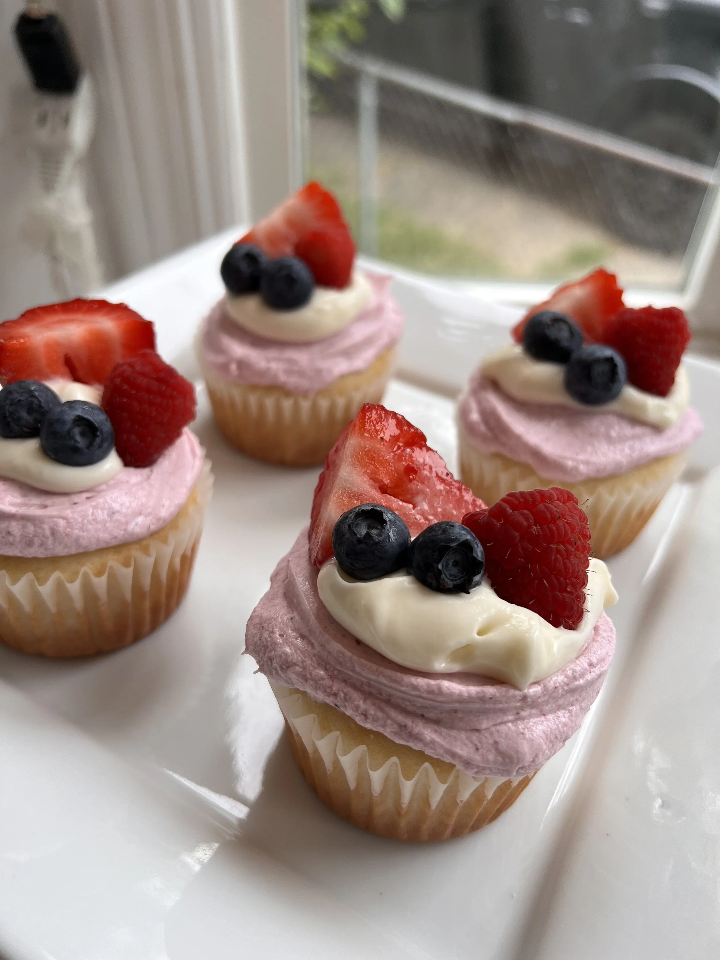 Four cupcakes with pink frosting, topped with strawberries, blueberries, and raspberries, placed on a white tray near a window.