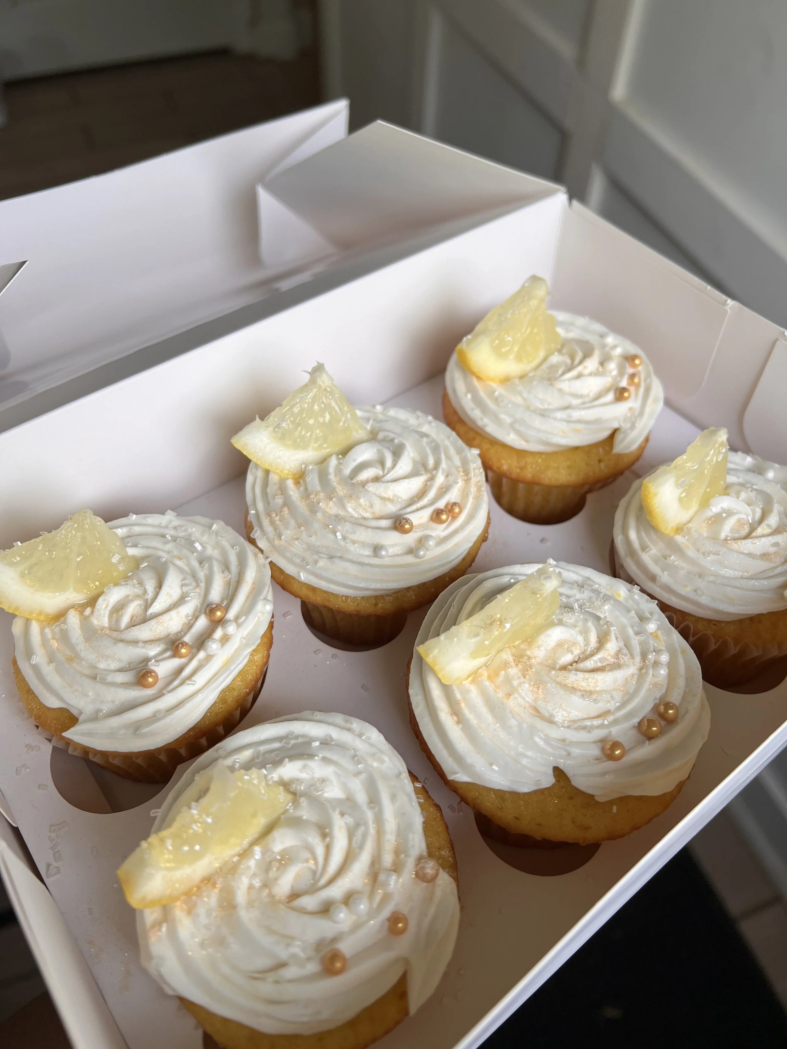 Six vanilla cupcakes with white frosting and decorative sprinkles in a white bakery box.