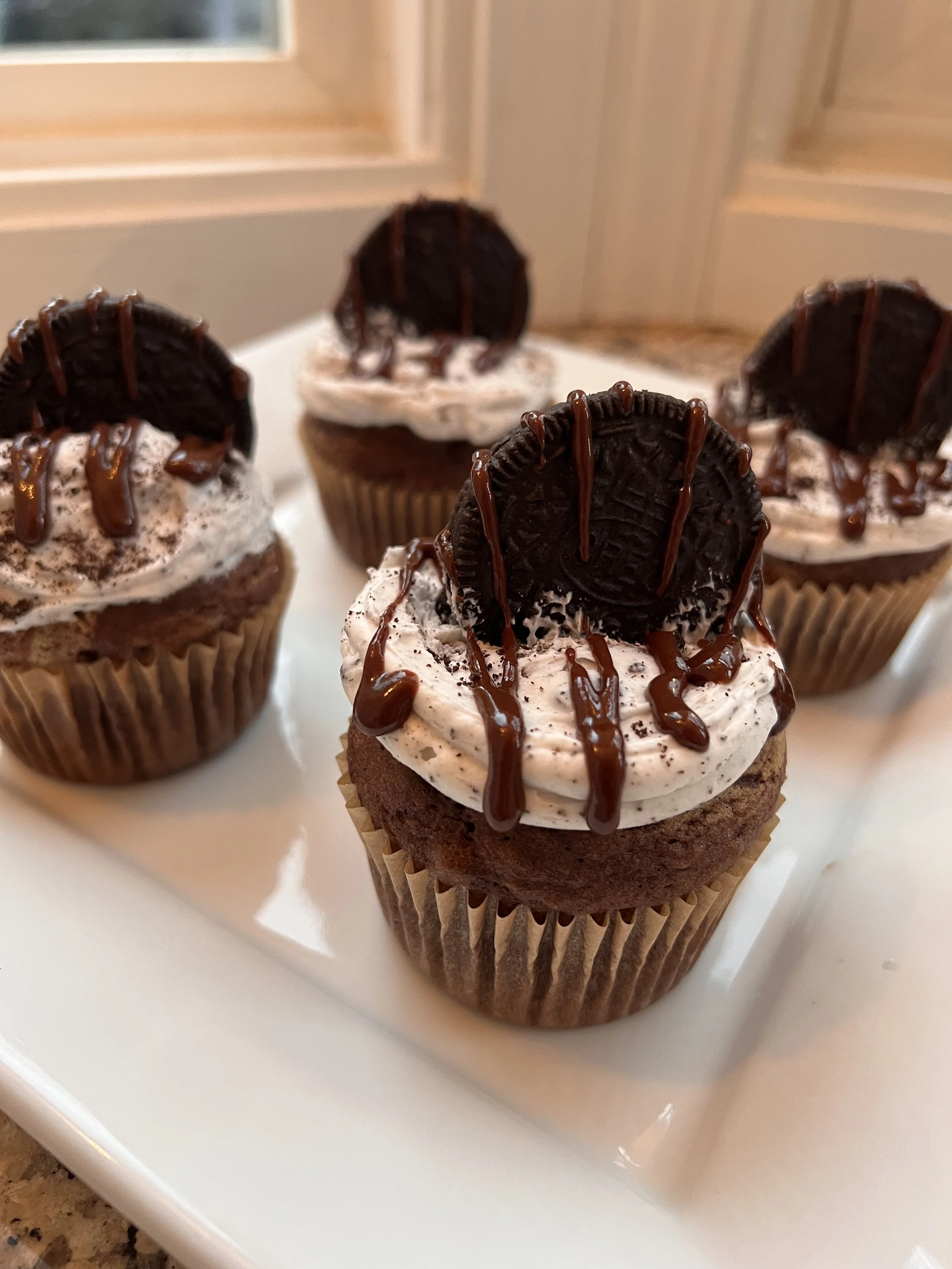 Chocolate cupcakes topped with Oreo cookies, chocolate drizzle, and cookies and cream frosting on a white tray.