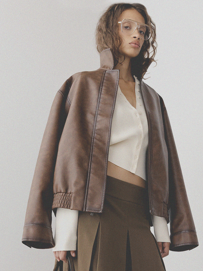 A woman with curly hair and glasses wearing a brown leather jacket over a white shirt and a brown skirt.