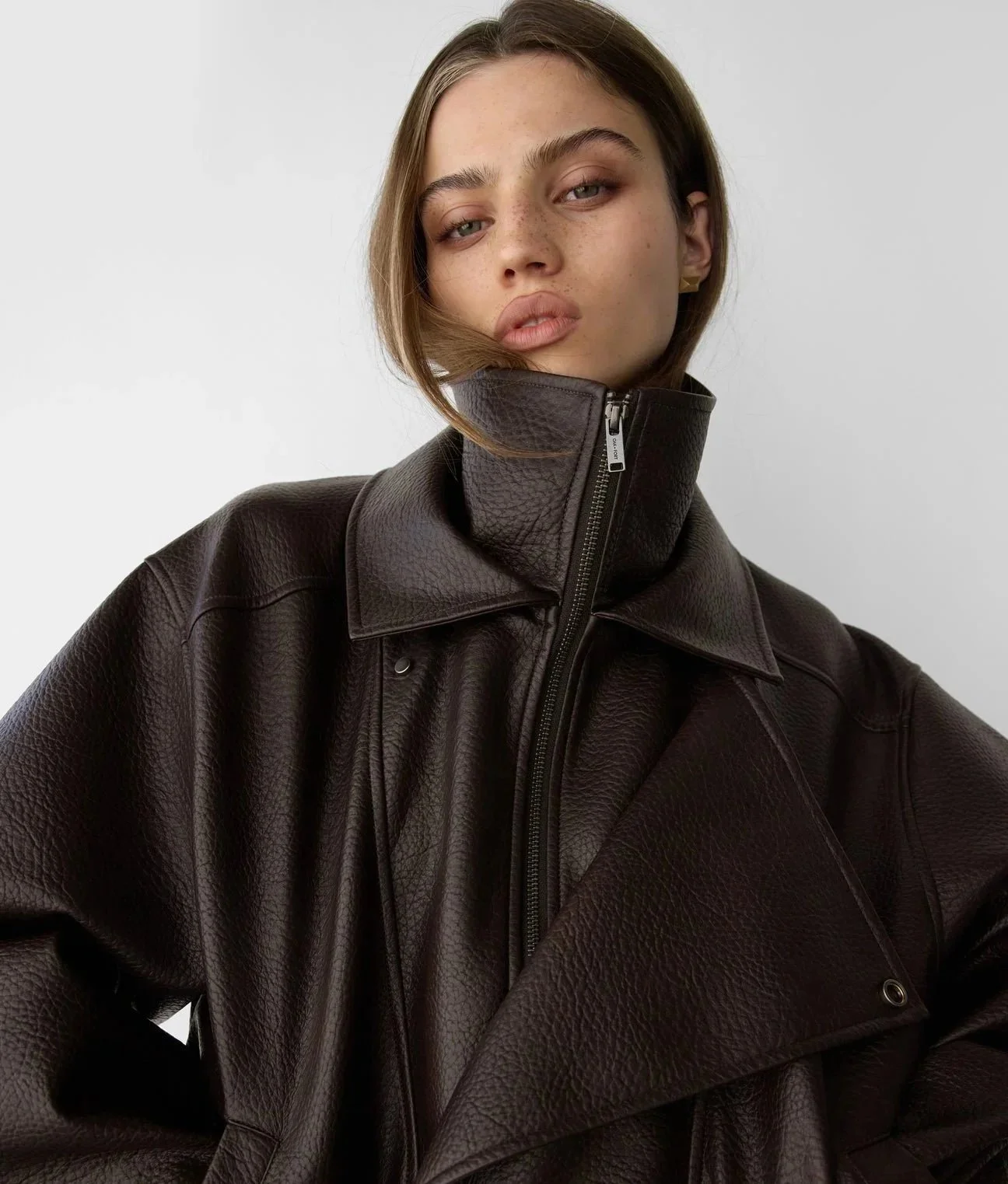 A young woman with light brown hair, freckles, and blue eyes wearing a dark brown leather jacket with a zipper, standing against a plain white background.