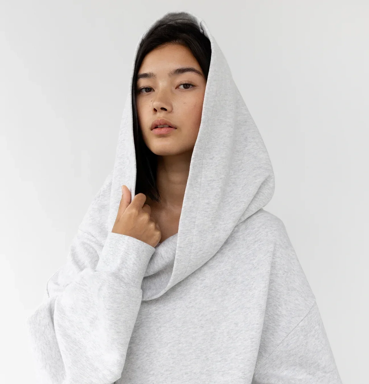 A woman with black hair and light skin wearing a light gray hoodie, holding the hood with her left hand, against a plain white background.
