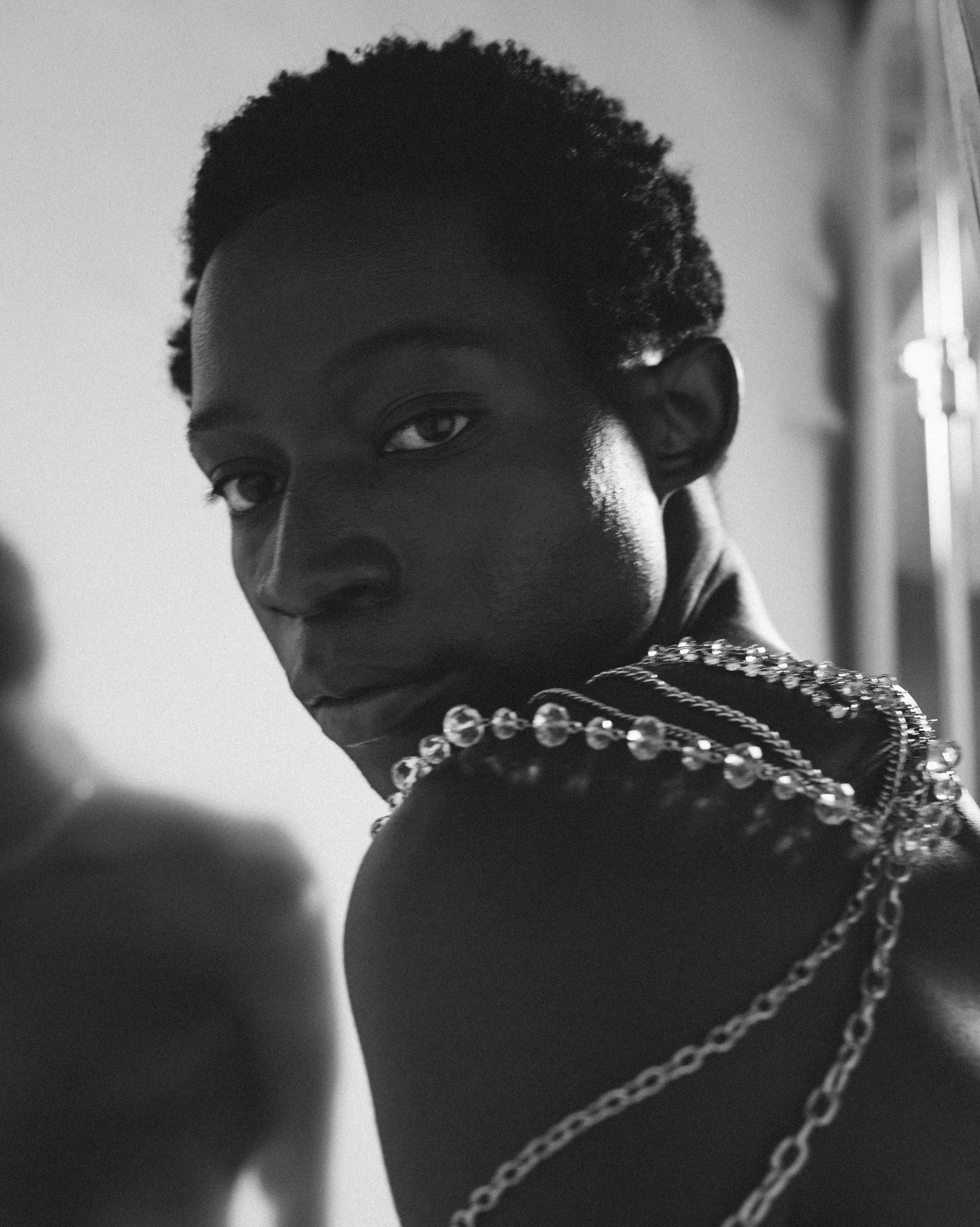 Close-up black and white photo of a person with short curly hair, covered partially by jewelry, in front of a window with light streaming in.