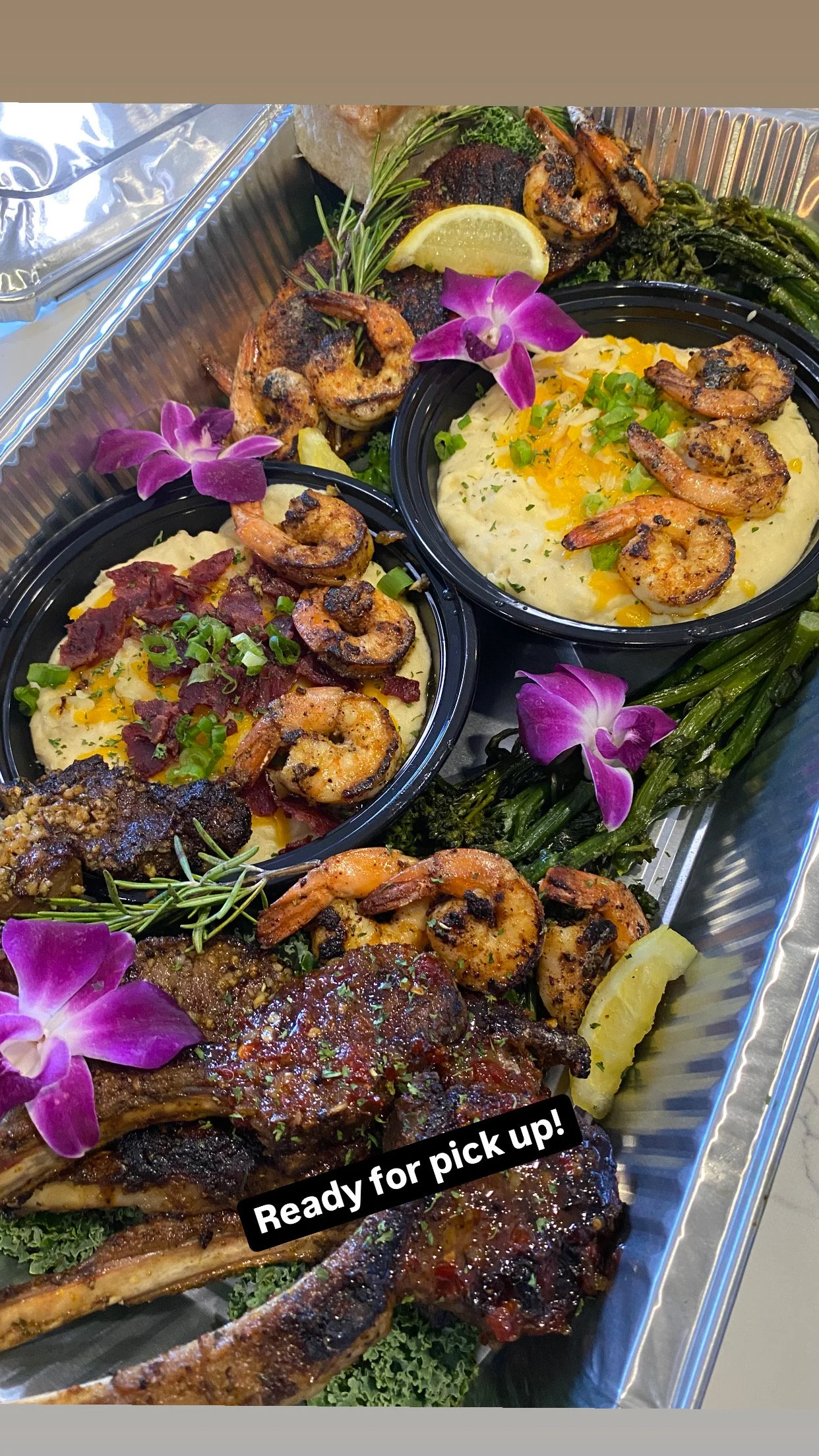 A tray of prepared food containing grilled shrimp, mashed potatoes, grilled vegetables, a piece of meat with tomato sauce, garnished with lemon slices, herbs, and purple orchids, with a black label reading "Ready for pick up!"