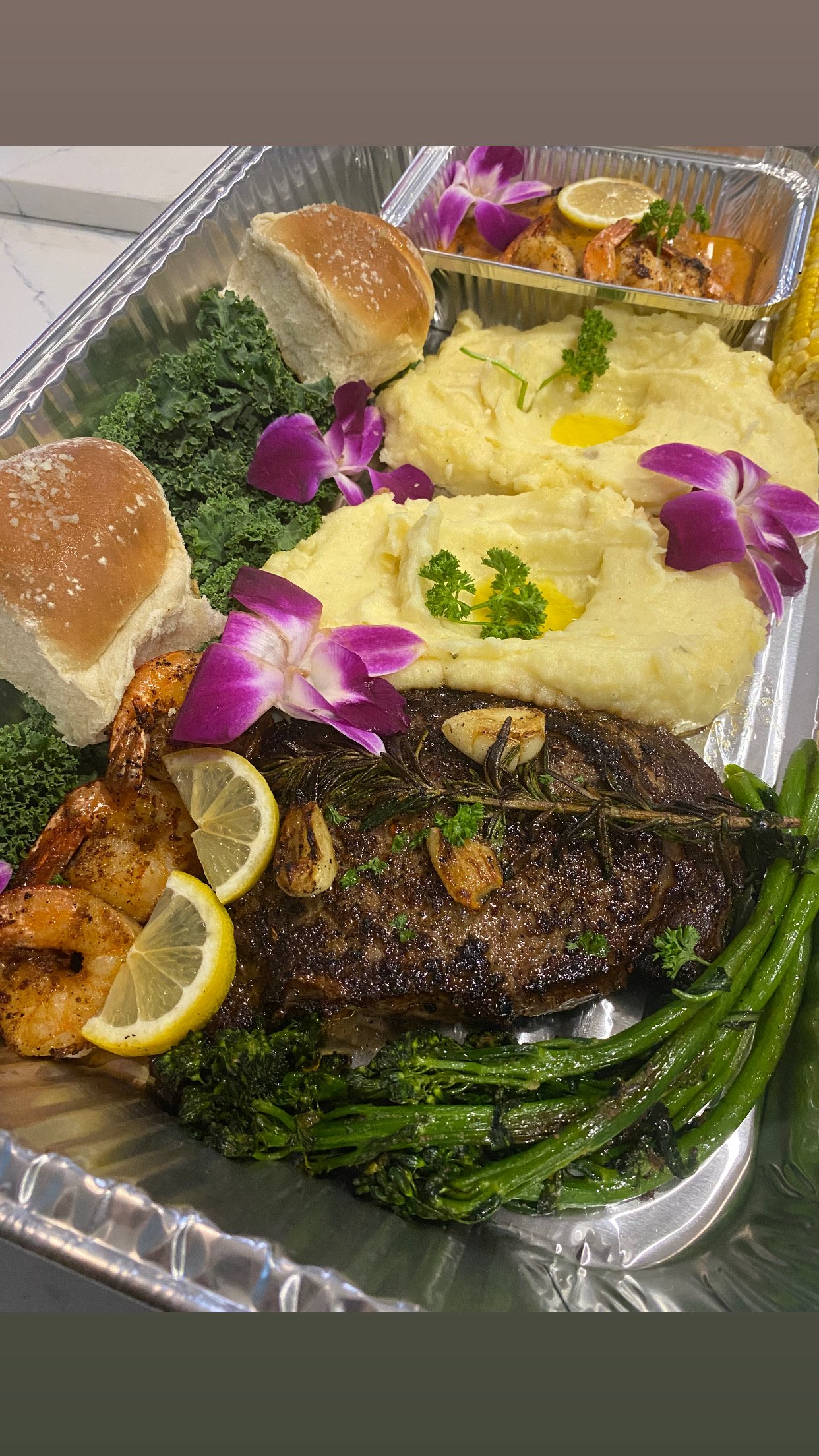 Aluminum tray with baked chicken, mashed potatoes with parsley and egg, garlic shrimp, green beans, grilled beef steak with lemon slices, corn on the cob, bread rolls, garlic and rosemary garnish, and edible purple orchid flowers.