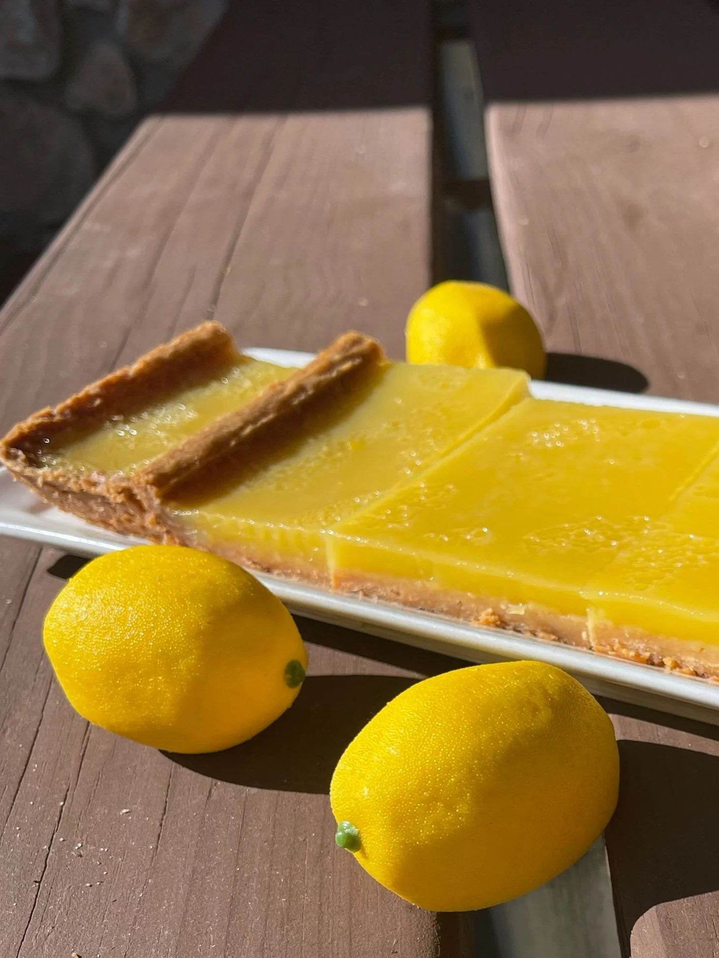Lemon tart with a graham cracker crust on a white rectangular plate, accompanied by three fresh lemons on a wooden outdoor table with sunlight and shadows.
