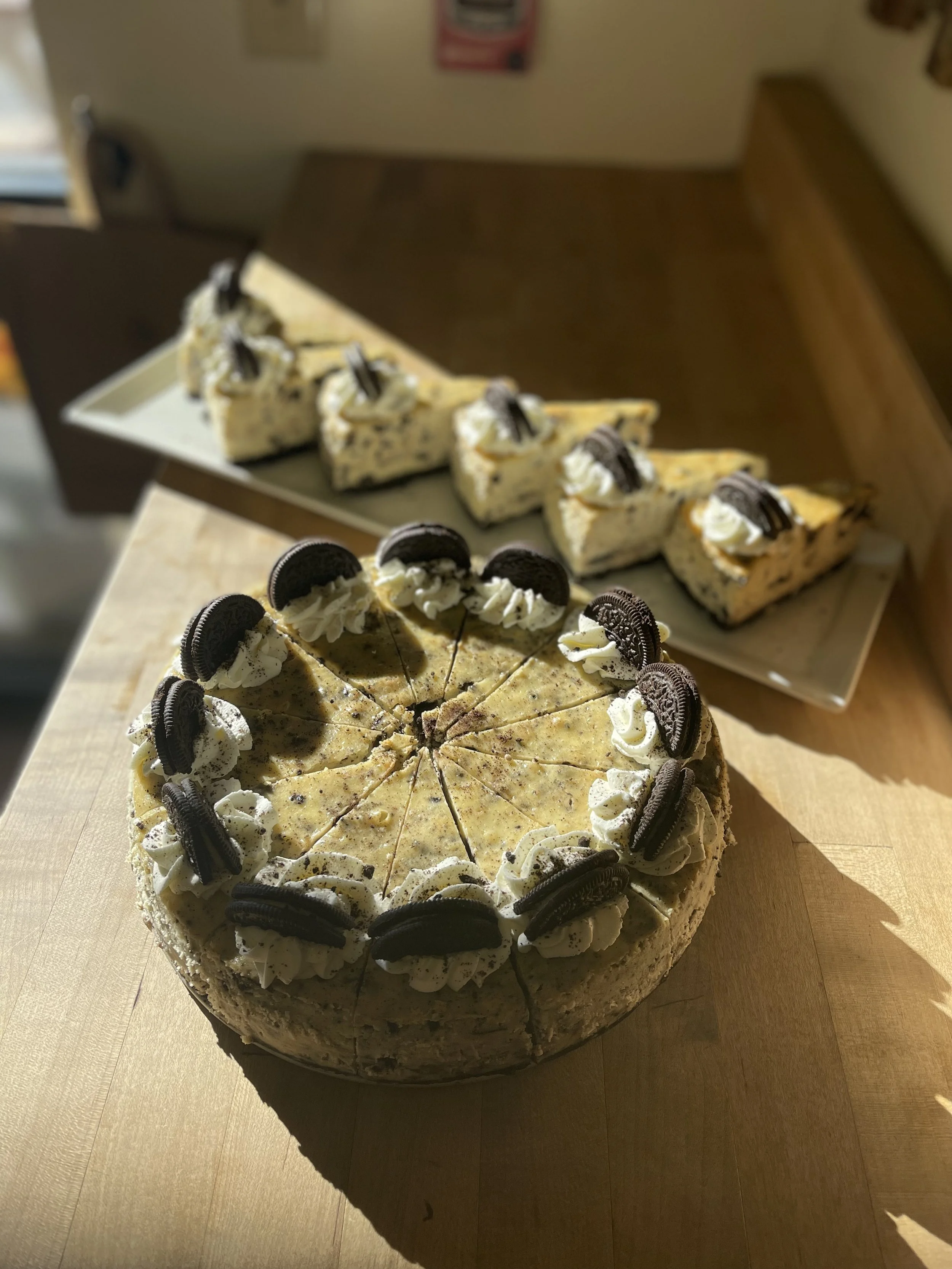 A large round cake decorated with Oreo cookies and whipped cream, with slices cut out. In the background, there are slices of cheesecake with Oreo toppings on a rectangular platter.