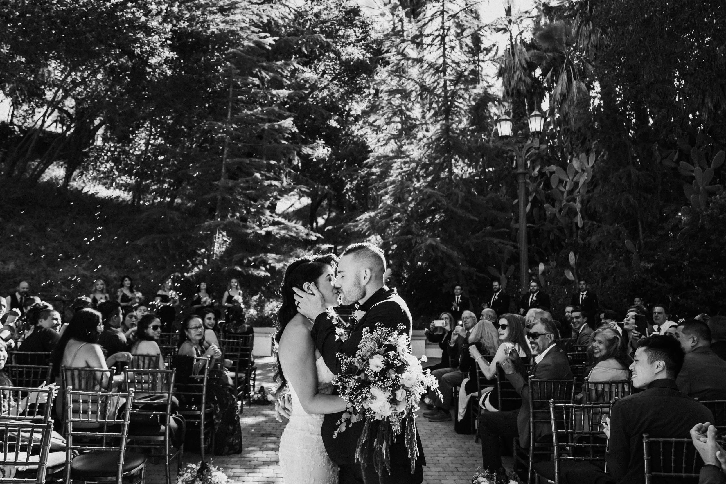 Wedding couple kissing outdoors, with seated guests applauding, surrounded by trees.