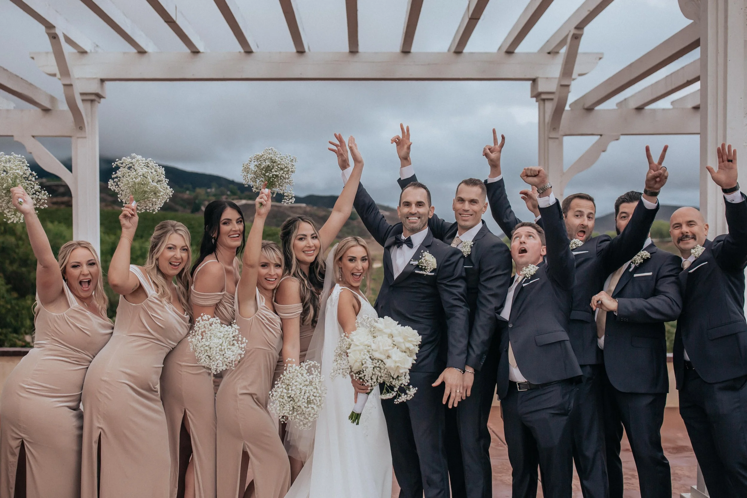 A joyful wedding party celebrating under a pergola, with bridesmaids in beige dresses holding flowers and groomsmen in dark suits.