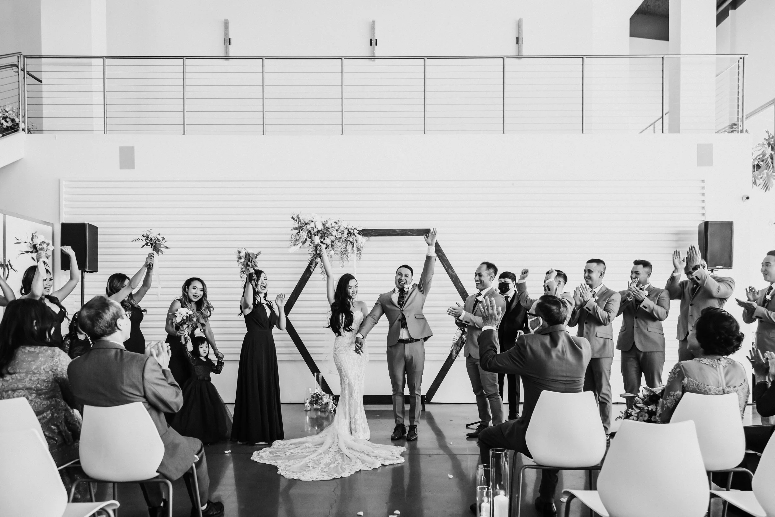 A wedding ceremony in a modern venue, showing a bride and groom joyfully celebrating in front of a geometric arch, surrounded by bridesmaids and groomsmen. Guests are seated, clapping and cheering.