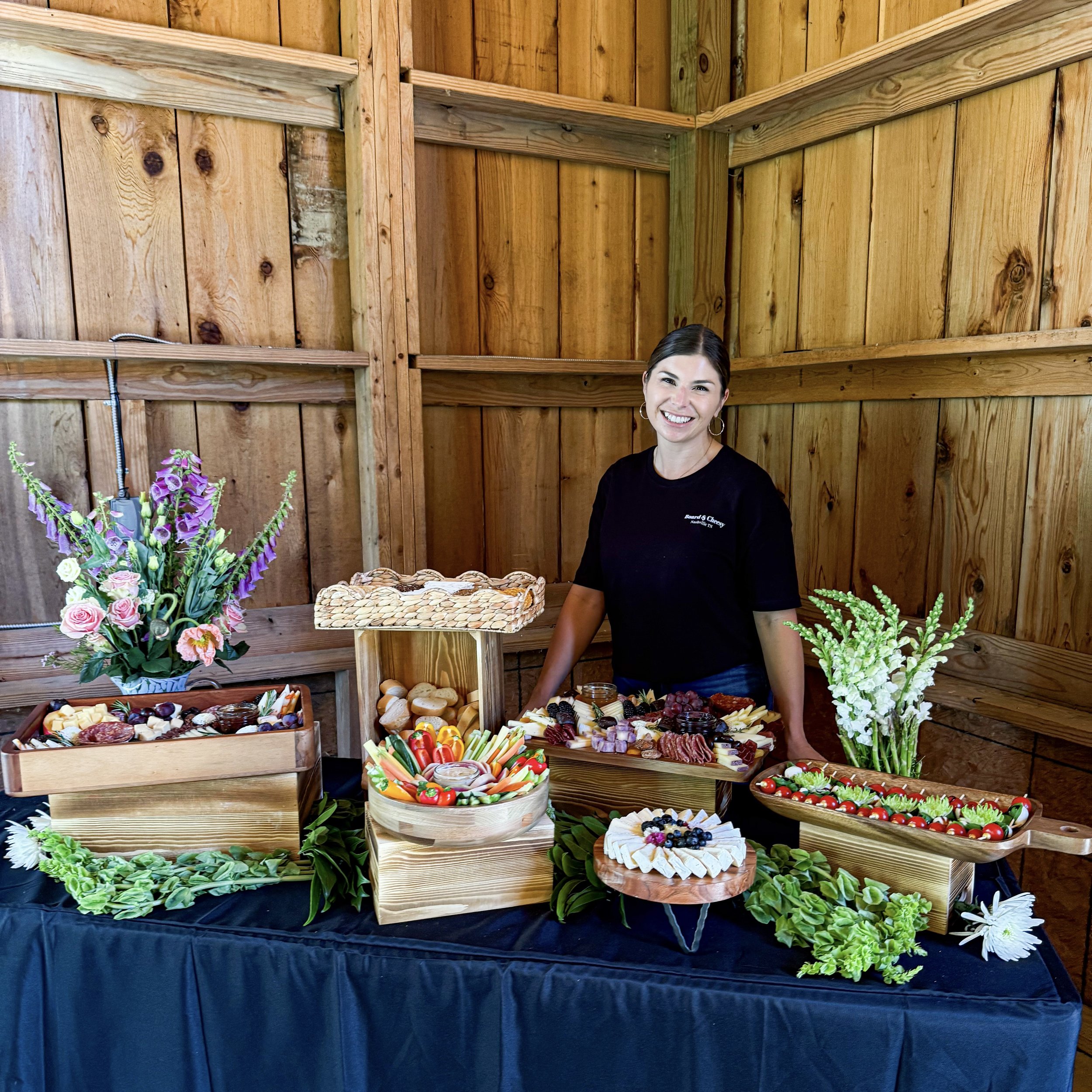 Woman standing at a table with a variety of cheeses, fruits, and vegetables, decorated with flowers and wooden elements, against wooden wall background.