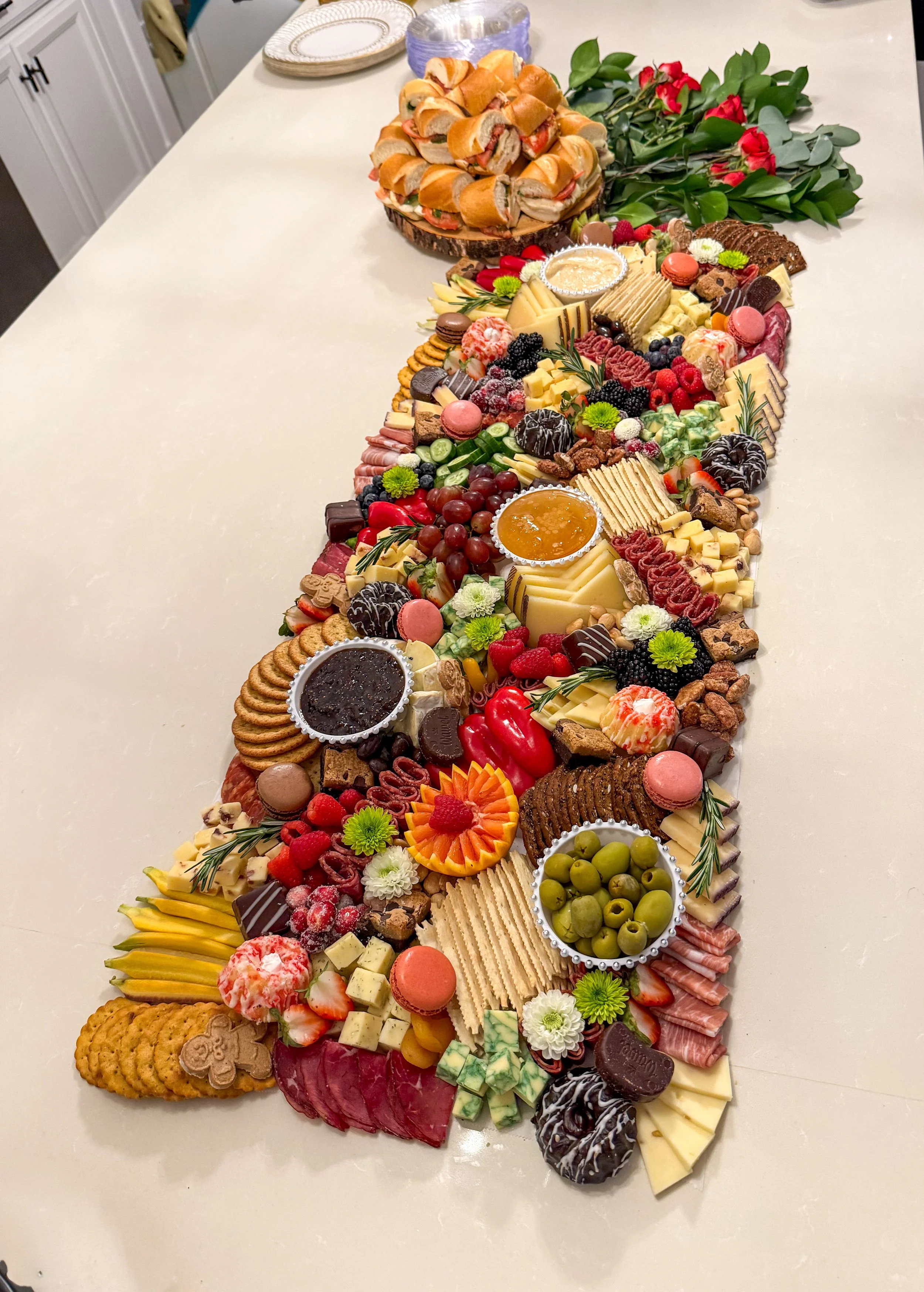 Grazing table with meats, cheeses, crackers, fruits, sweets, olives, vegetables, jam, honey, hummus, and small sandwiches outlined with greenery and red roses. 