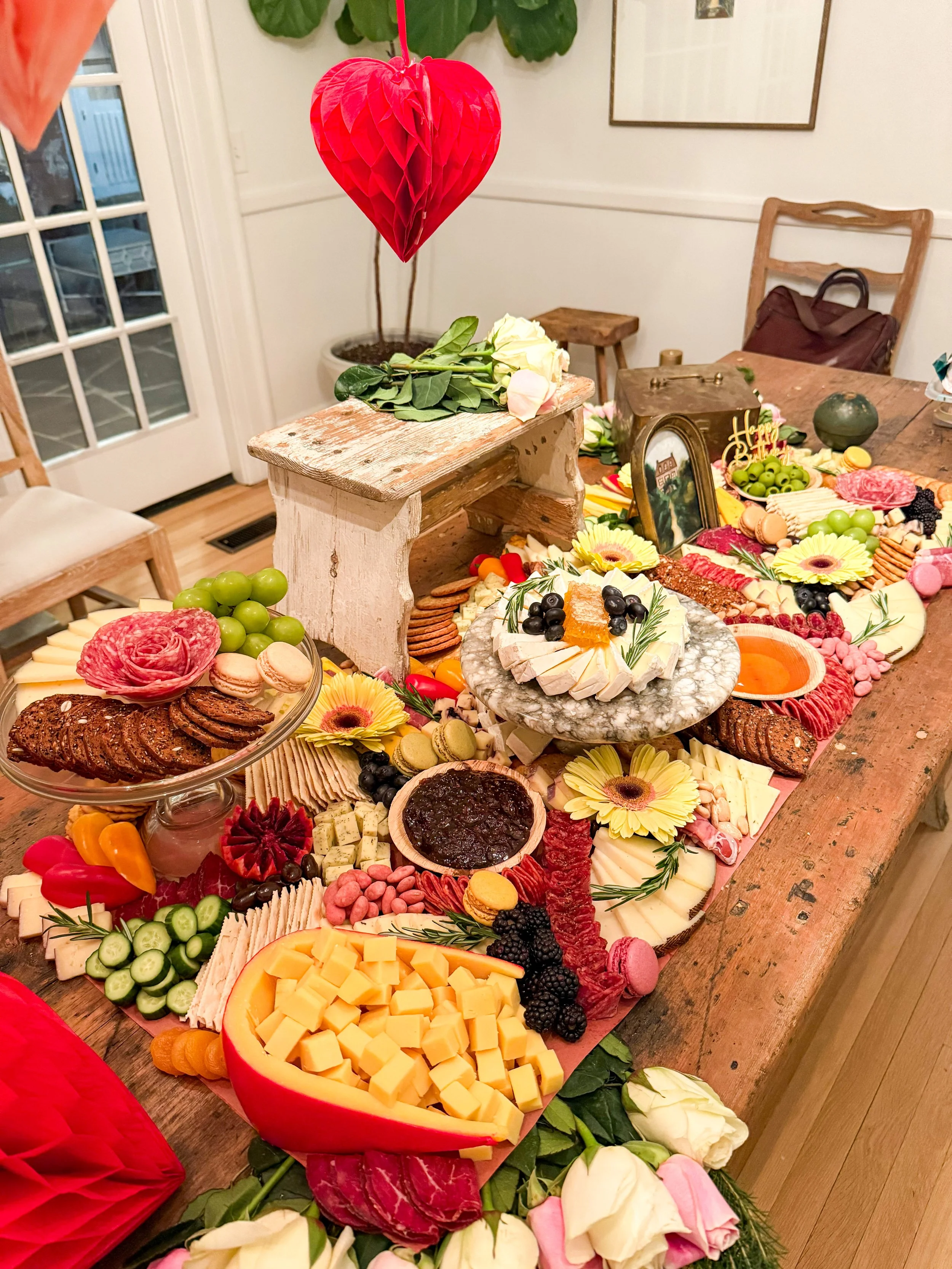 Multi dimensional grazing table with meats, cheeses, crackers, fresh fruit, honey and jam with colorful roses outlining it.