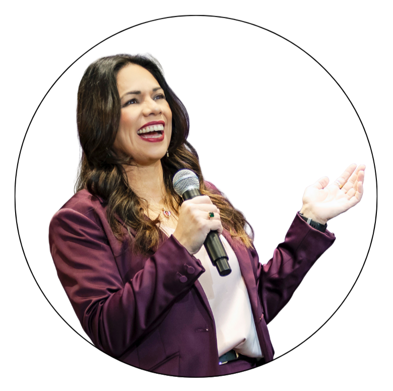 Esme Graff with long brown hair, wearing a maroon blazer and white blouse, smiling while holding a microphone and gesturing with her other hand.
