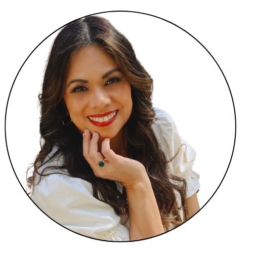 Esme, with long wavy brown hair, wearing a white top, smiling and resting her chin on her hand, against a white background.