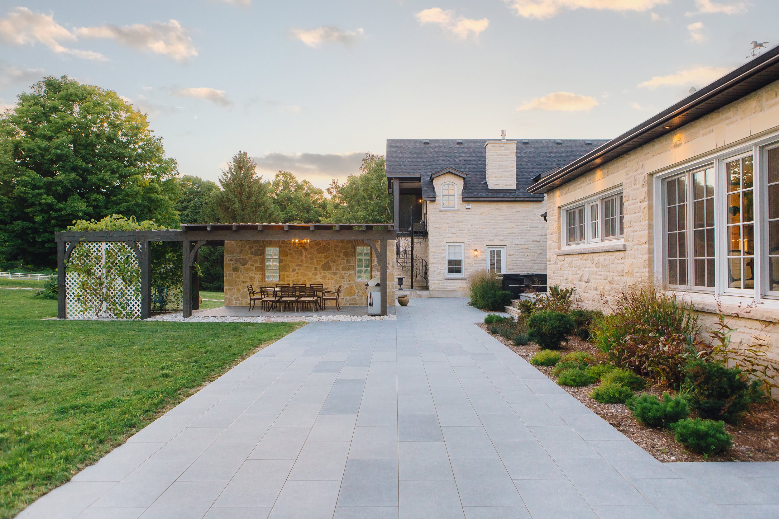 Outdoor patio with porcelain pavers, adjacent to a pool, surrounded by a Guillotined armour stone retaining wall with shrubs and ornamental grasses.