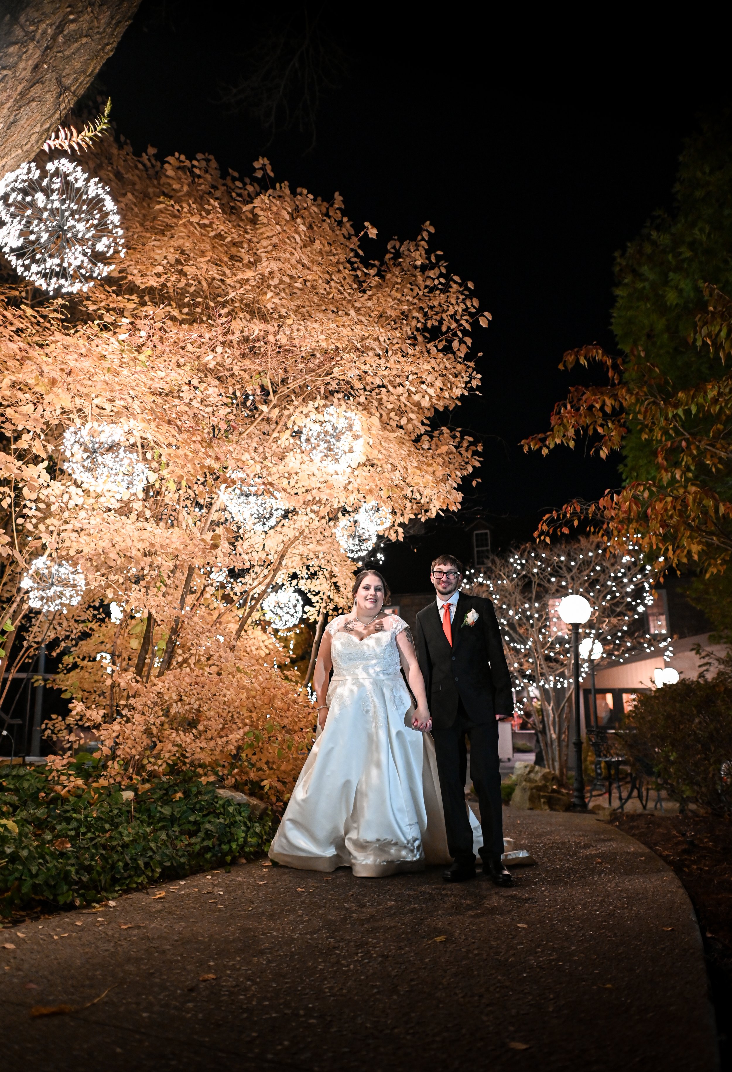 Bride and groom holding hands standing on a pathway at night, surrounded by illuminated trees with autumn leaves and decorative lights.