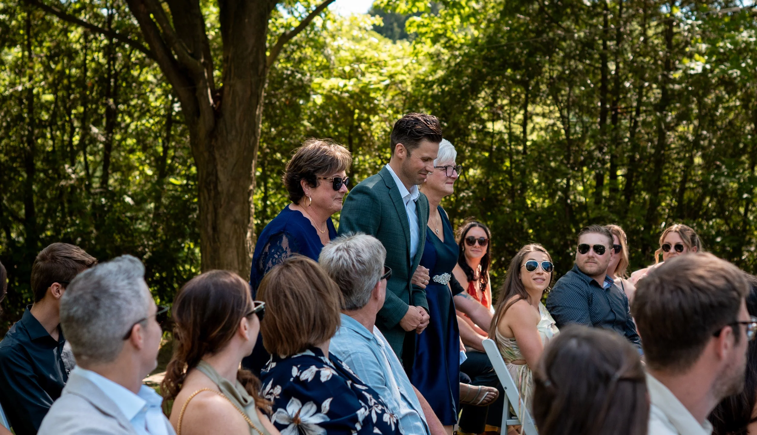 People attending an outdoor event during the daytime, seated on white chairs in a wooded area with green trees in the background.