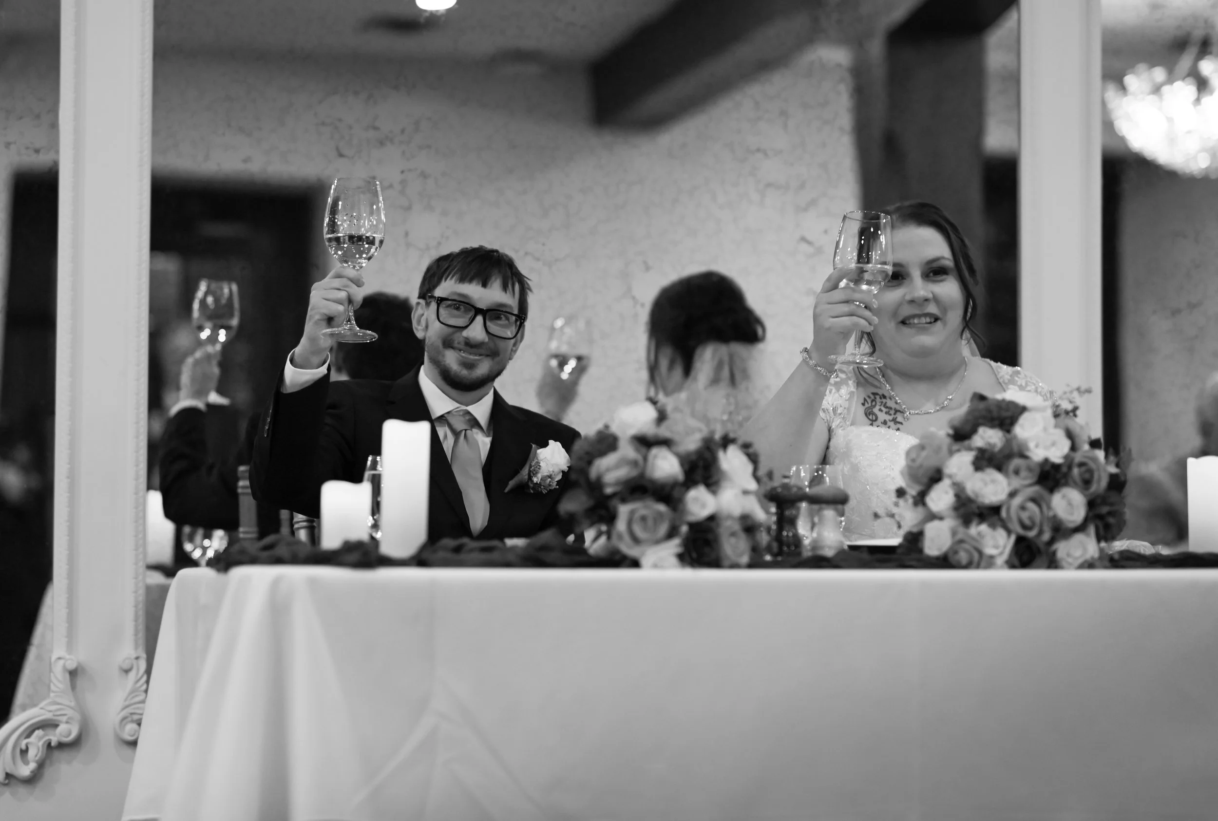 A bride and groom at their wedding reception sitting at a decorated table with wine glasses raised, surrounded by floral arrangements and candles, with a mirror reflecting their image.