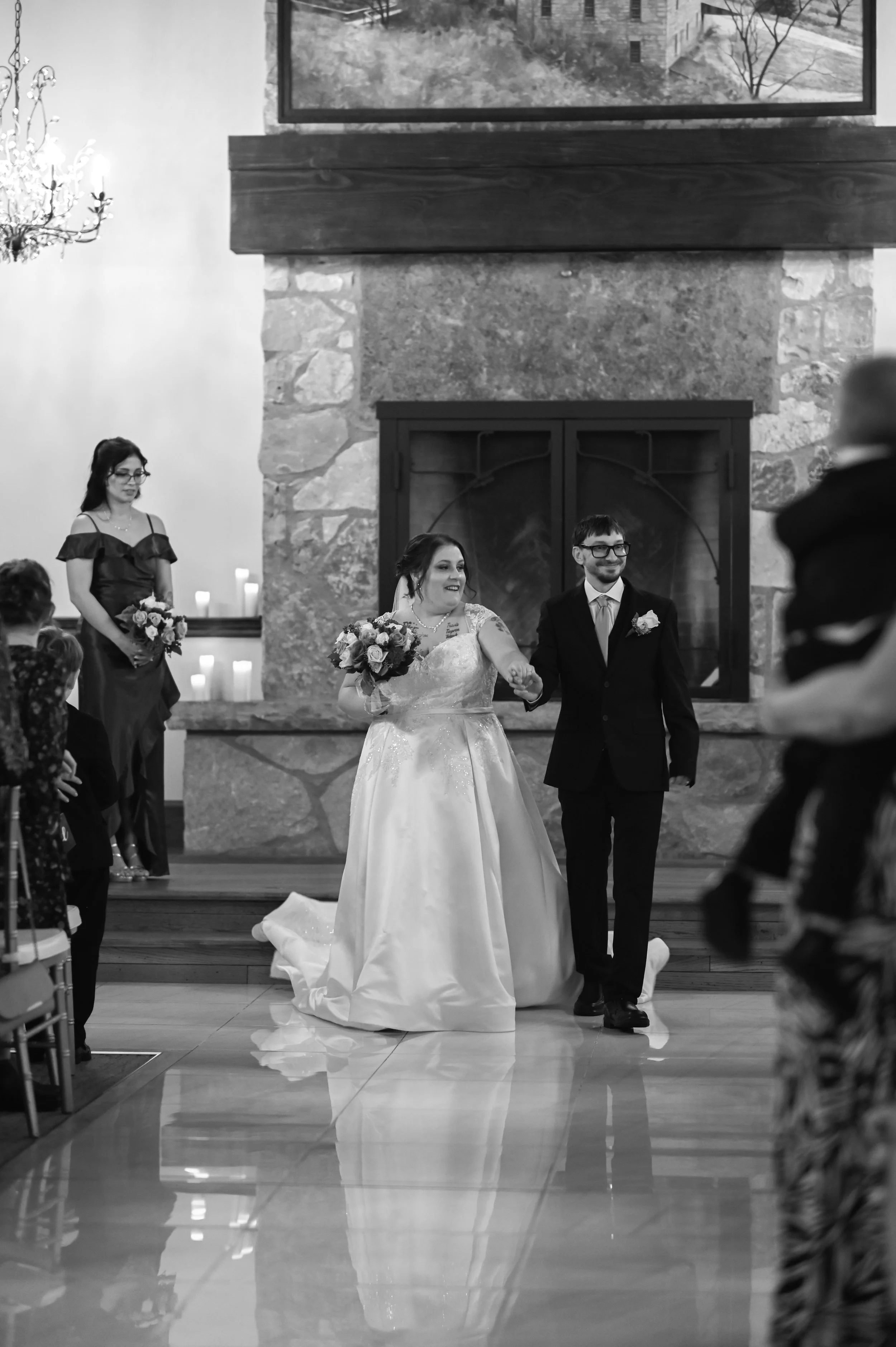 A black and white photo of a wedding ceremony with a bride and groom walking hand in hand in front of a large stone fireplace, surrounded by guests.