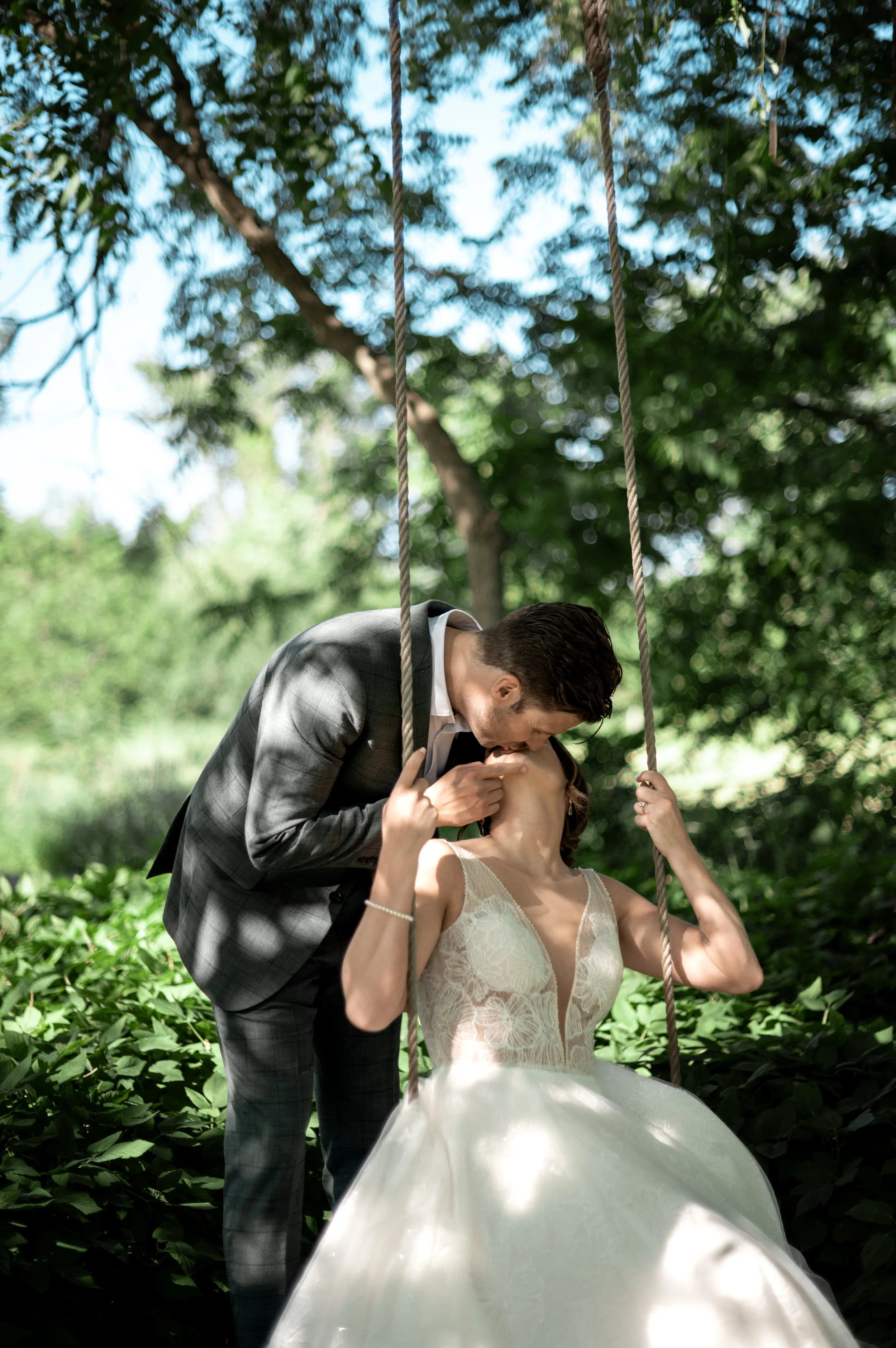 A couple dressed in wedding attire sharing a kiss on a swing outdoors, surrounded by green foliage and beneath a tree canopy.