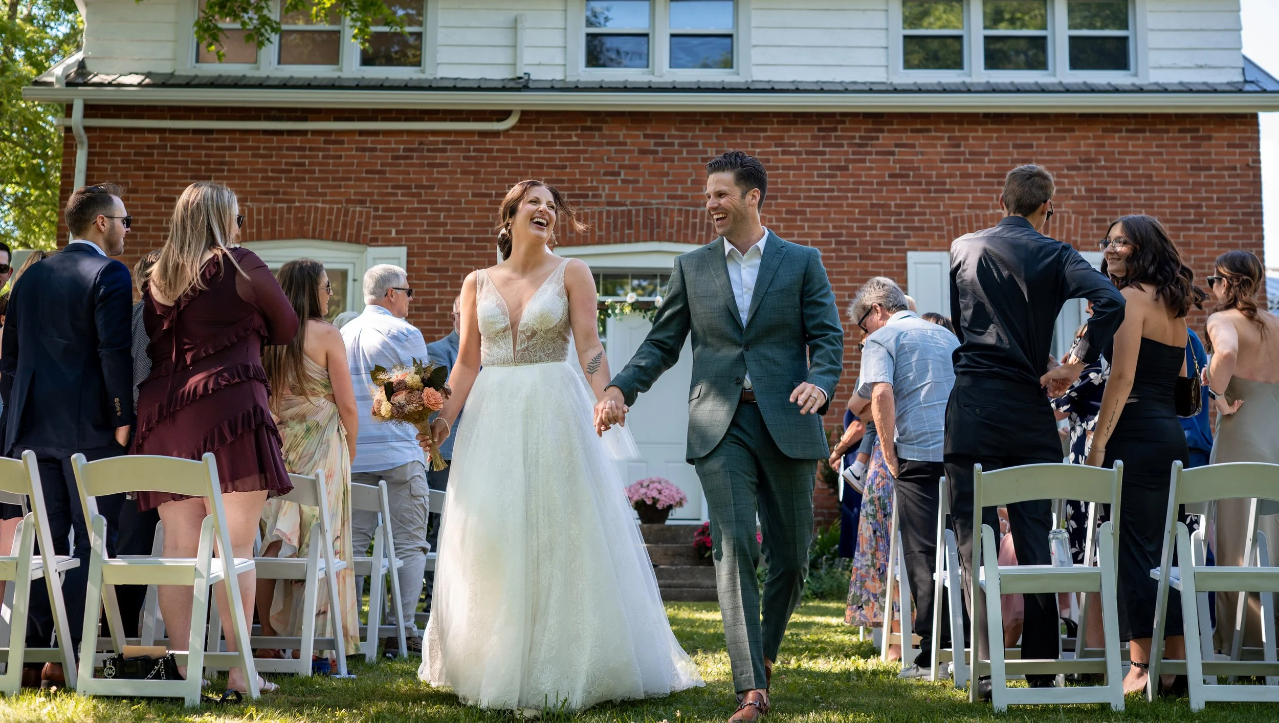 A bride and groom celebrating at an outdoor wedding ceremony. The bride is wearing a white dress and holding a bouquet, while the groom is in a green suit. They are smiling and holding hands, surrounded by guests in colorful attire.