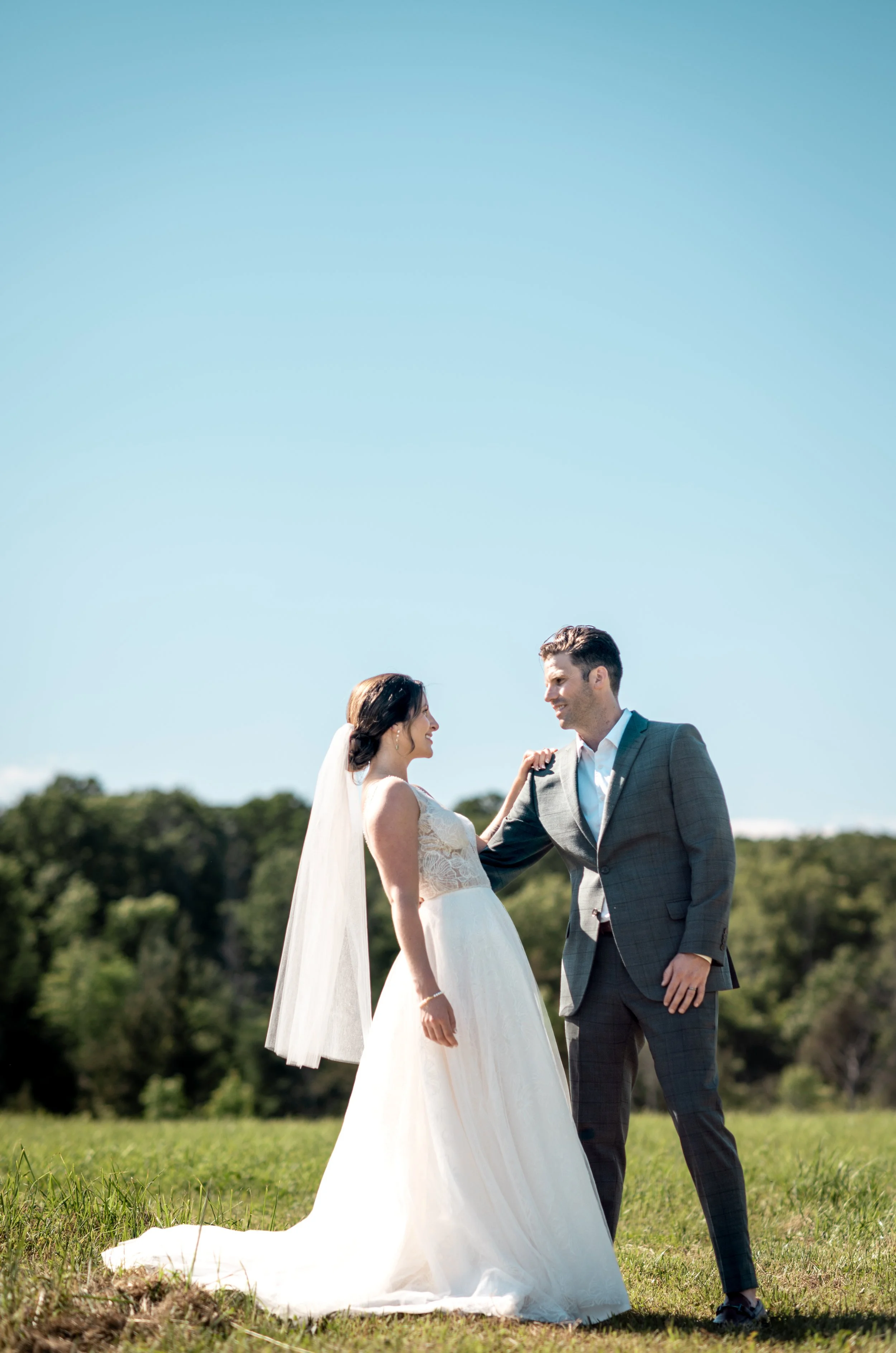A bride and groom are standing outdoors on grass, smiling and looking at each other, with a background of trees and a clear blue sky.
