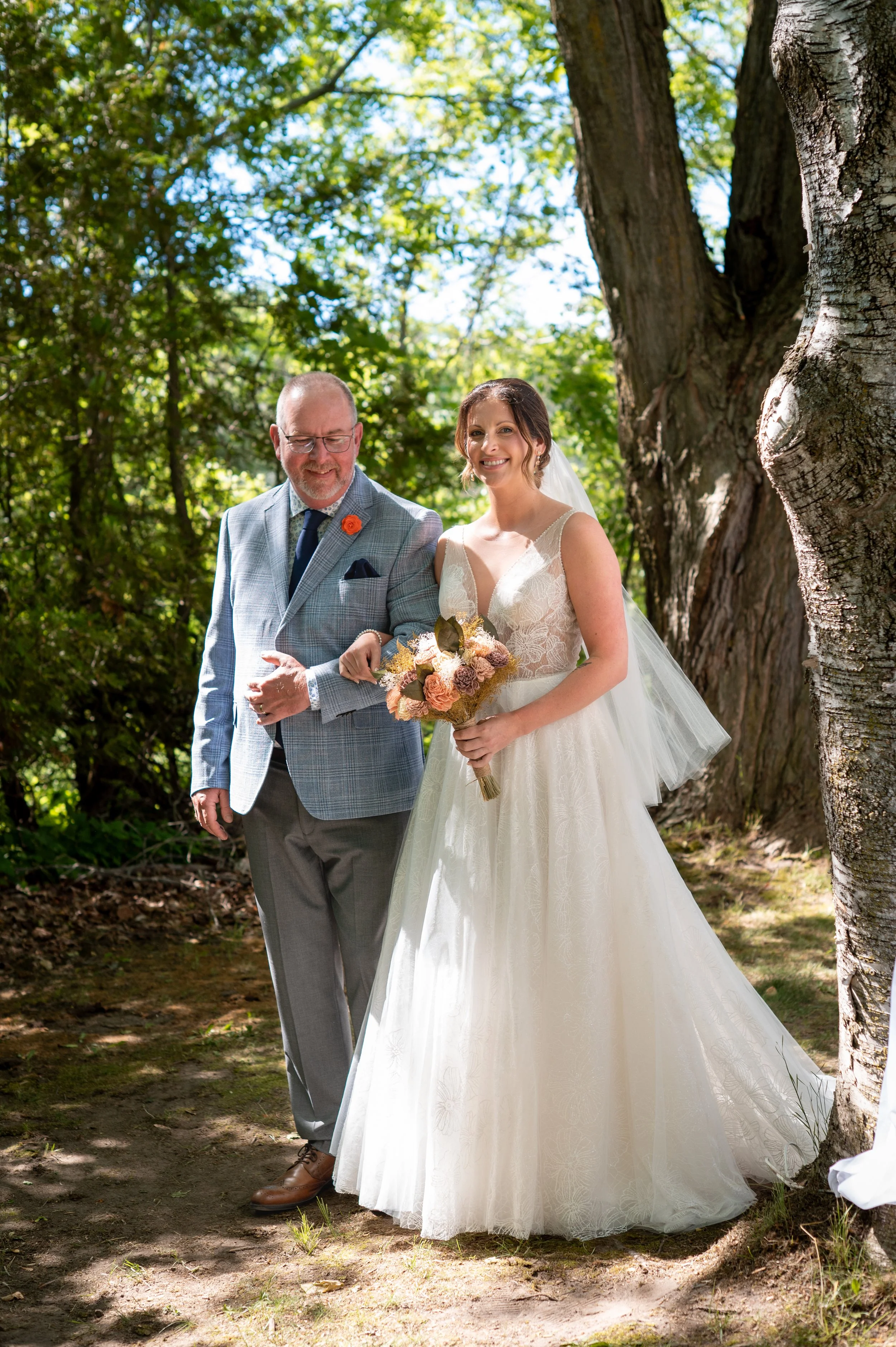 A bride in a white wedding dress holding a bouquet of flowers, walking outdoors with a man in a light blue checked suit. They are surrounded by green trees and sunlight filtering through leaves.