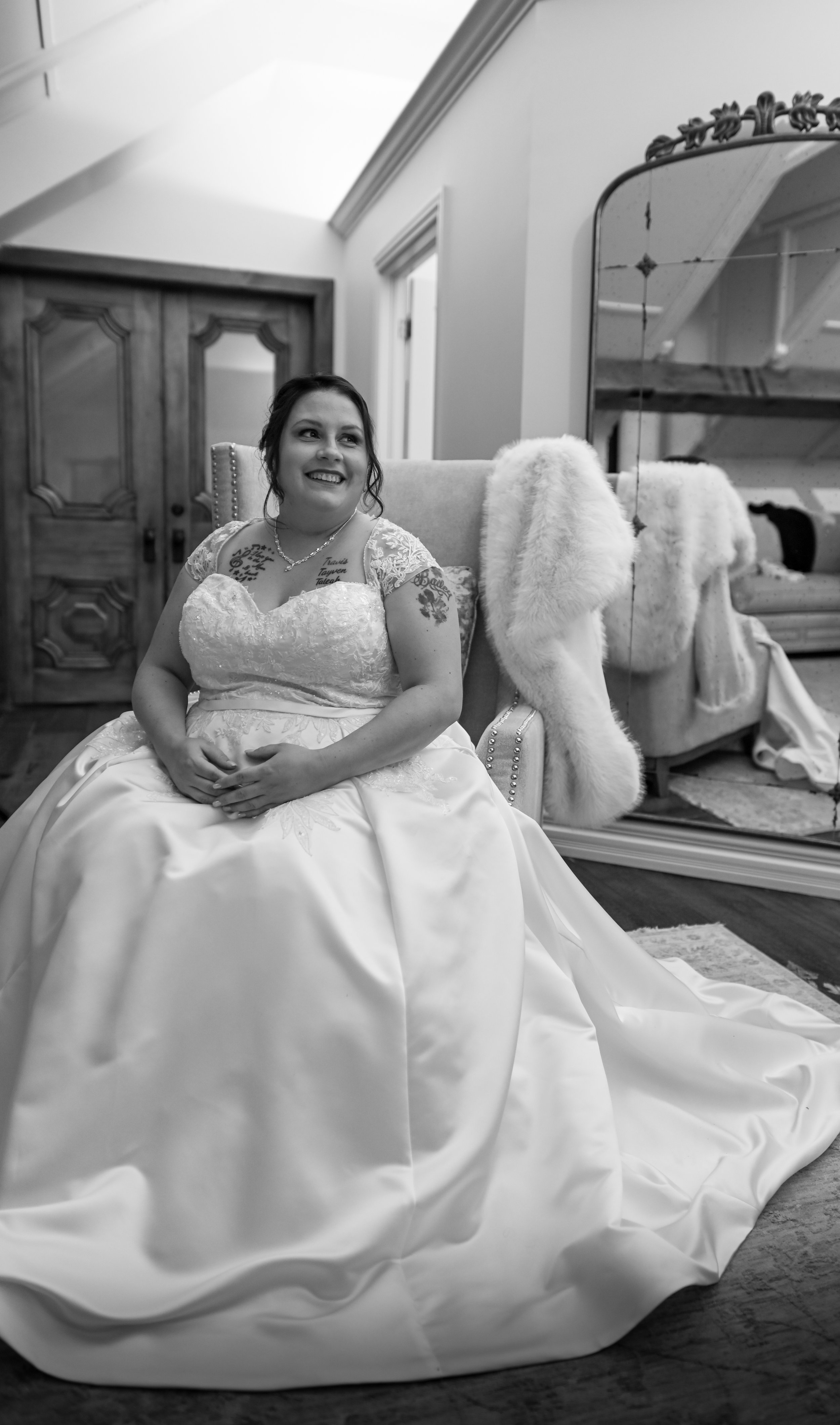 A smiling woman in a wedding dress sitting on a chair in a room with a large mirror and a furry blanket.