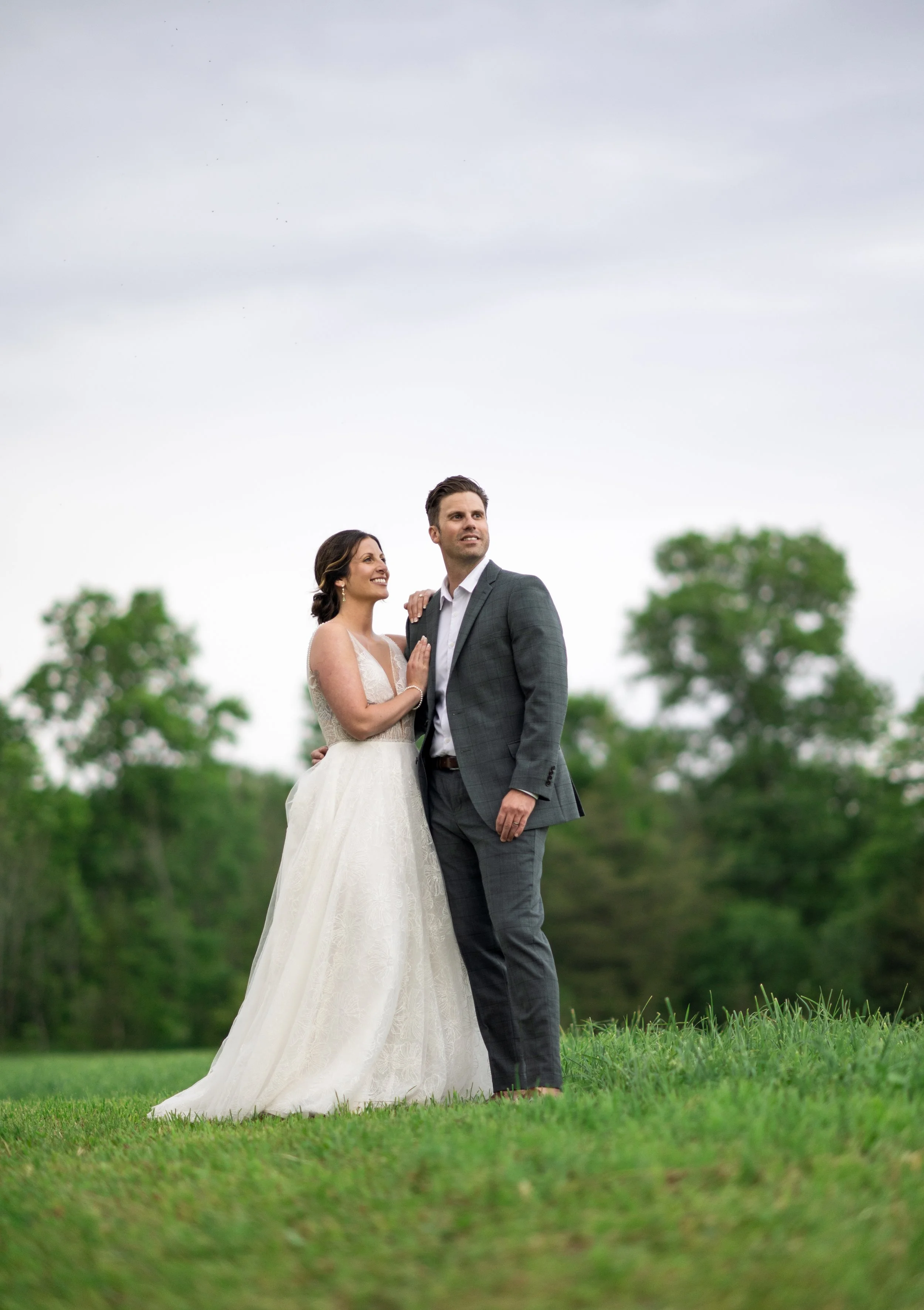 A newlywed couple standing in a grassy field with trees in the background, the bride in a strapless white gown smiling up at the groom, who is wearing a gray suit, white shirt, and no tie, both looking towards the distance on a cloudy day.