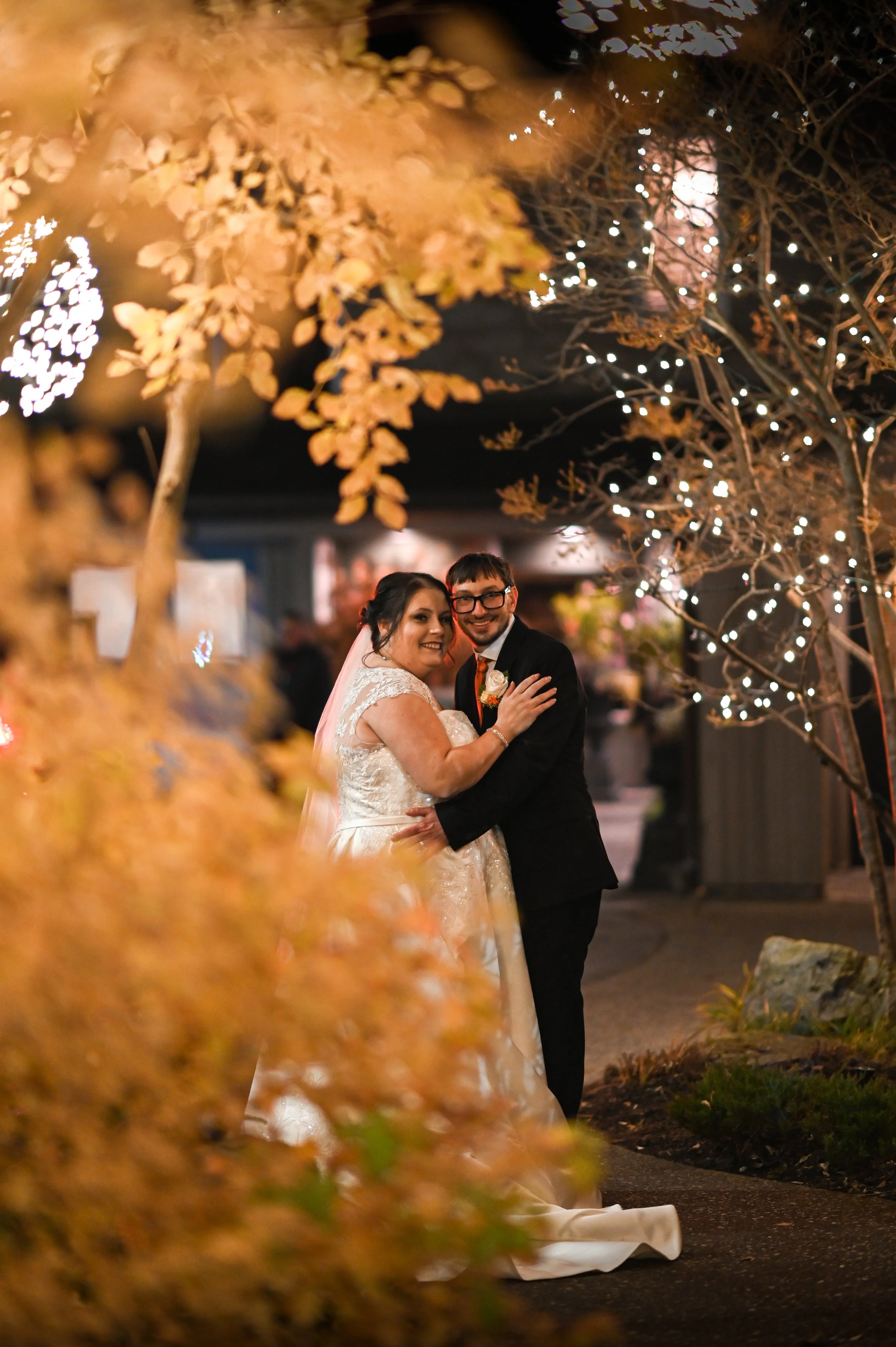 A bride and groom smiling and embracing at night, surrounded by trees decorated with string lights and autumn-colored foliage.