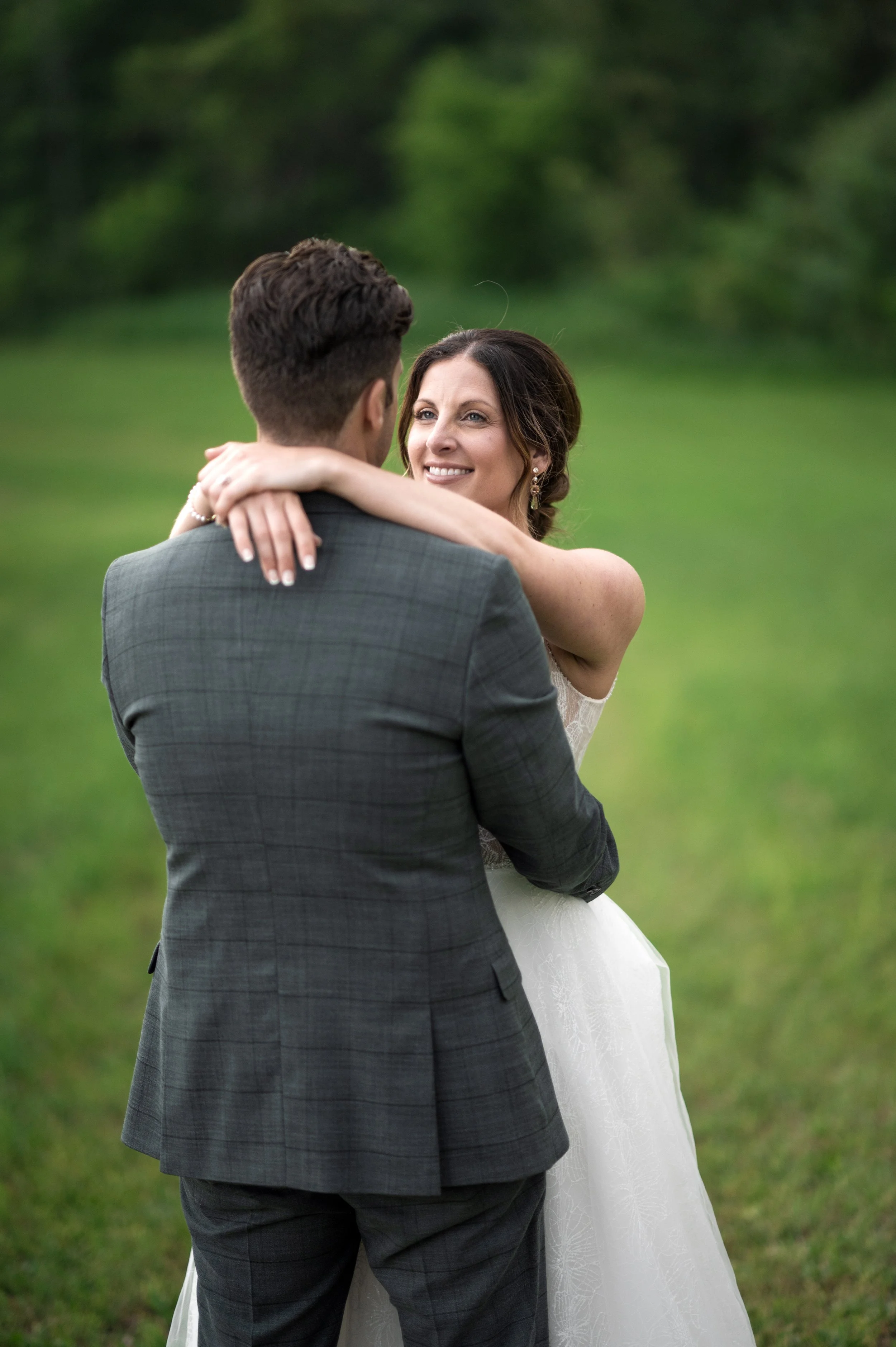 A bride and groom embracing outdoors on a grassy area with trees in the background.