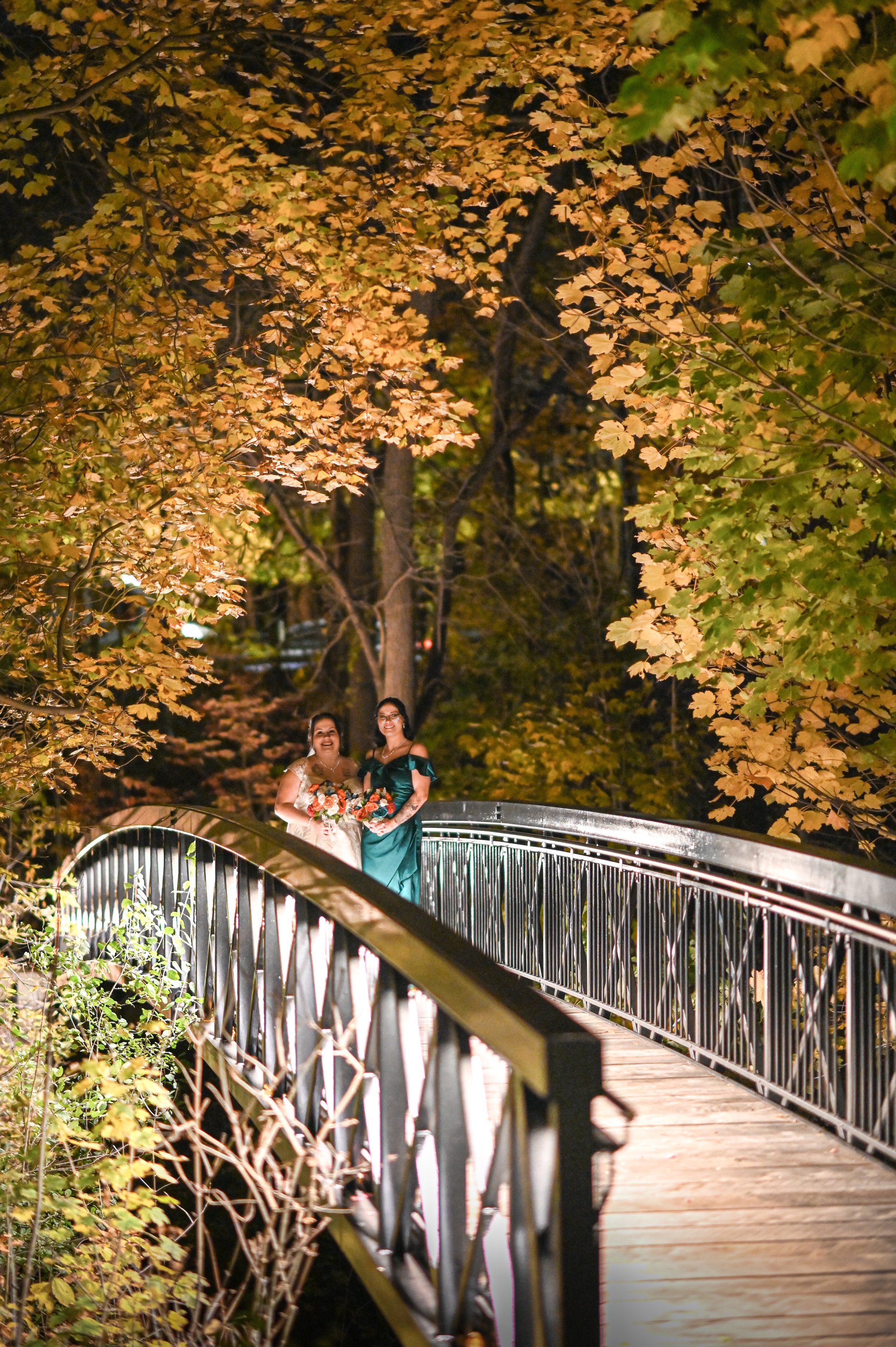 Two women in formal dresses holding bouquets stand on a lit, outdoor bridge surrounded by autumn-colored trees at night.