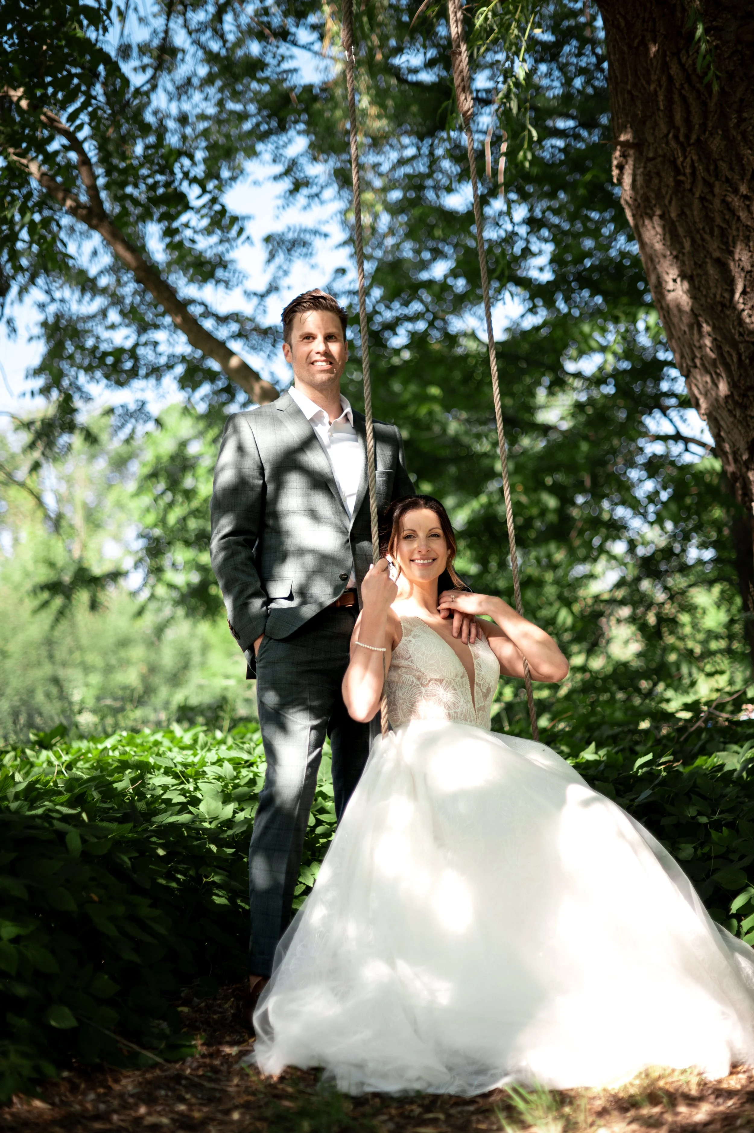 A young couple dressed in wedding attire outdoors on a swing hanging from a tree, surrounded by green foliage and sunlight filtering through the leaves.
