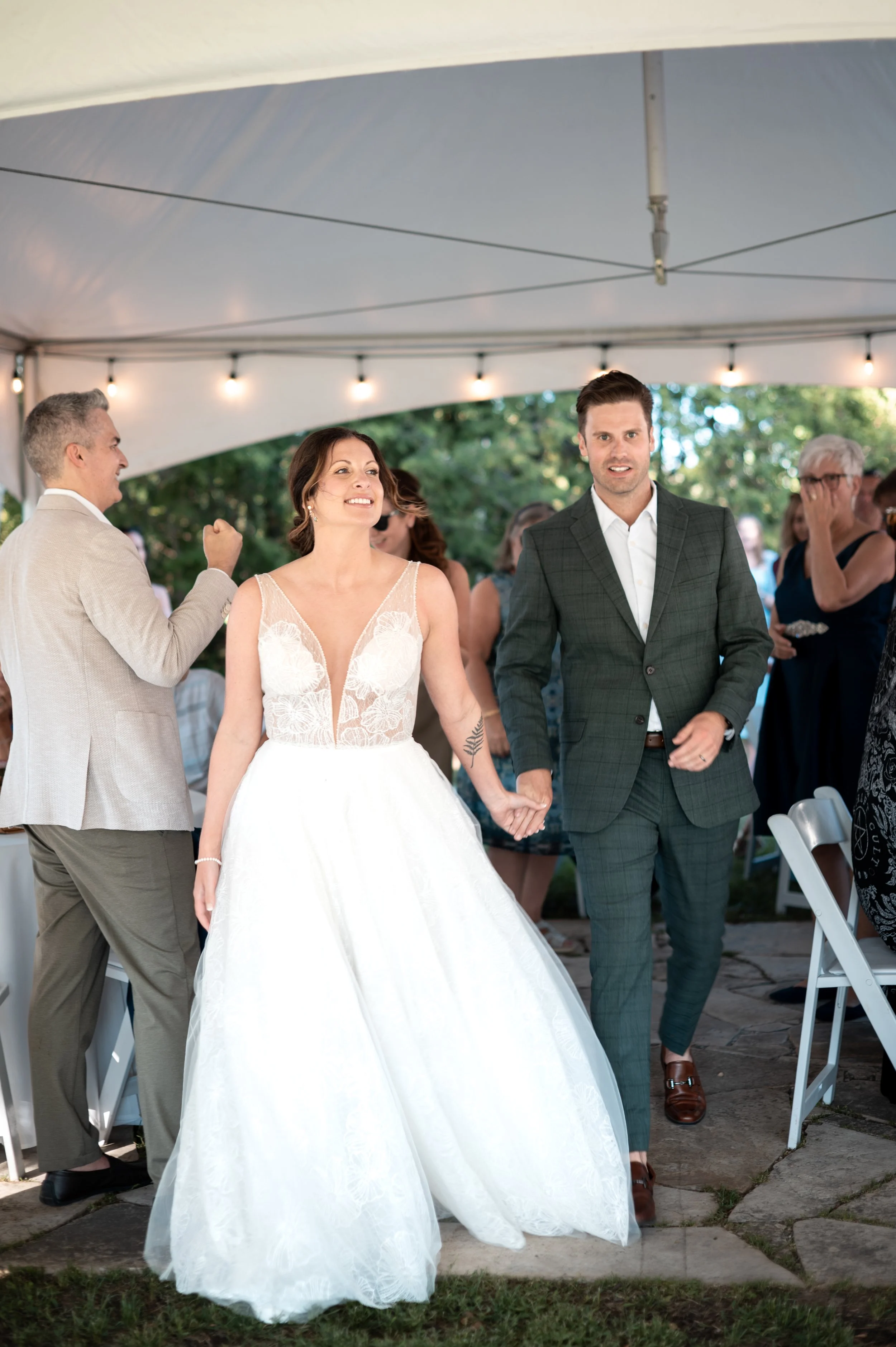 A bride and groom are holding hands and walking together during their wedding celebration under a white canopy with string lights. The bride is wearing a white wedding dress with a lace bodice and the groom is wearing a dark gray suit with a white sh