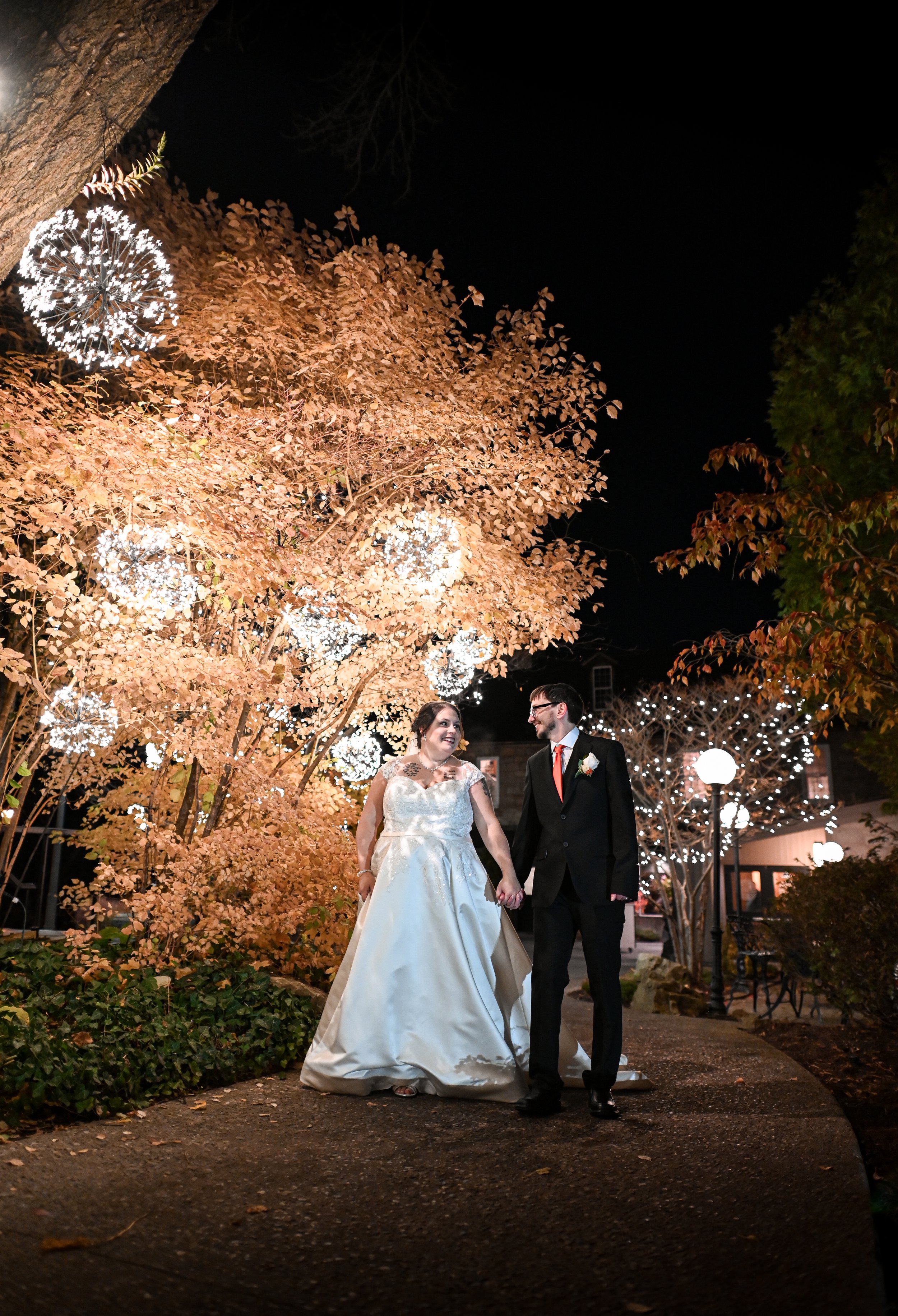 Bride and groom holding hands walking on a pathway at night, surrounded by illuminated trees and decorative lights.