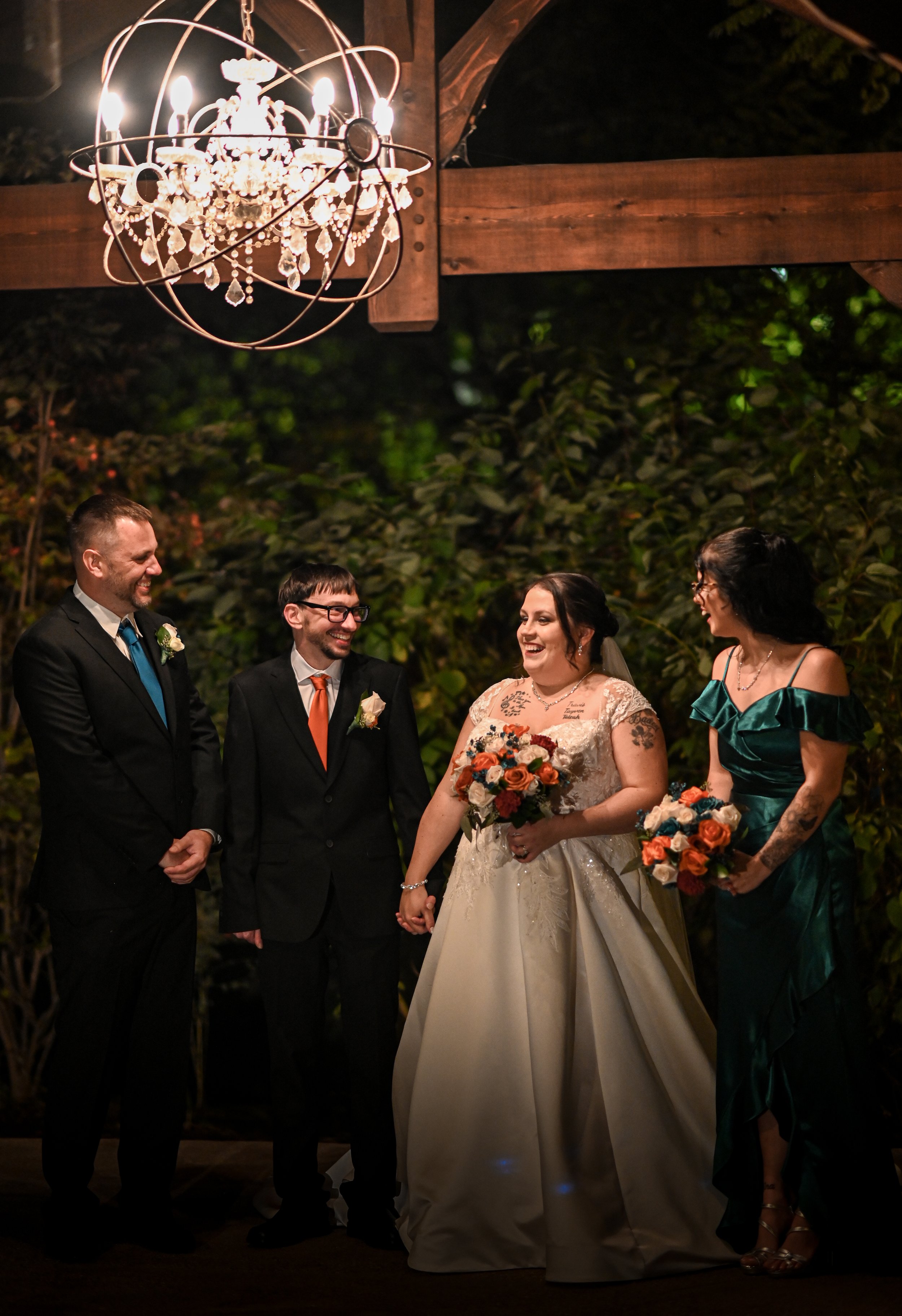 A wedding ceremony with a bride in a white gown holding a bouquet, a groom in a black suit, and two women in gowns, standing outdoors under a chandelier